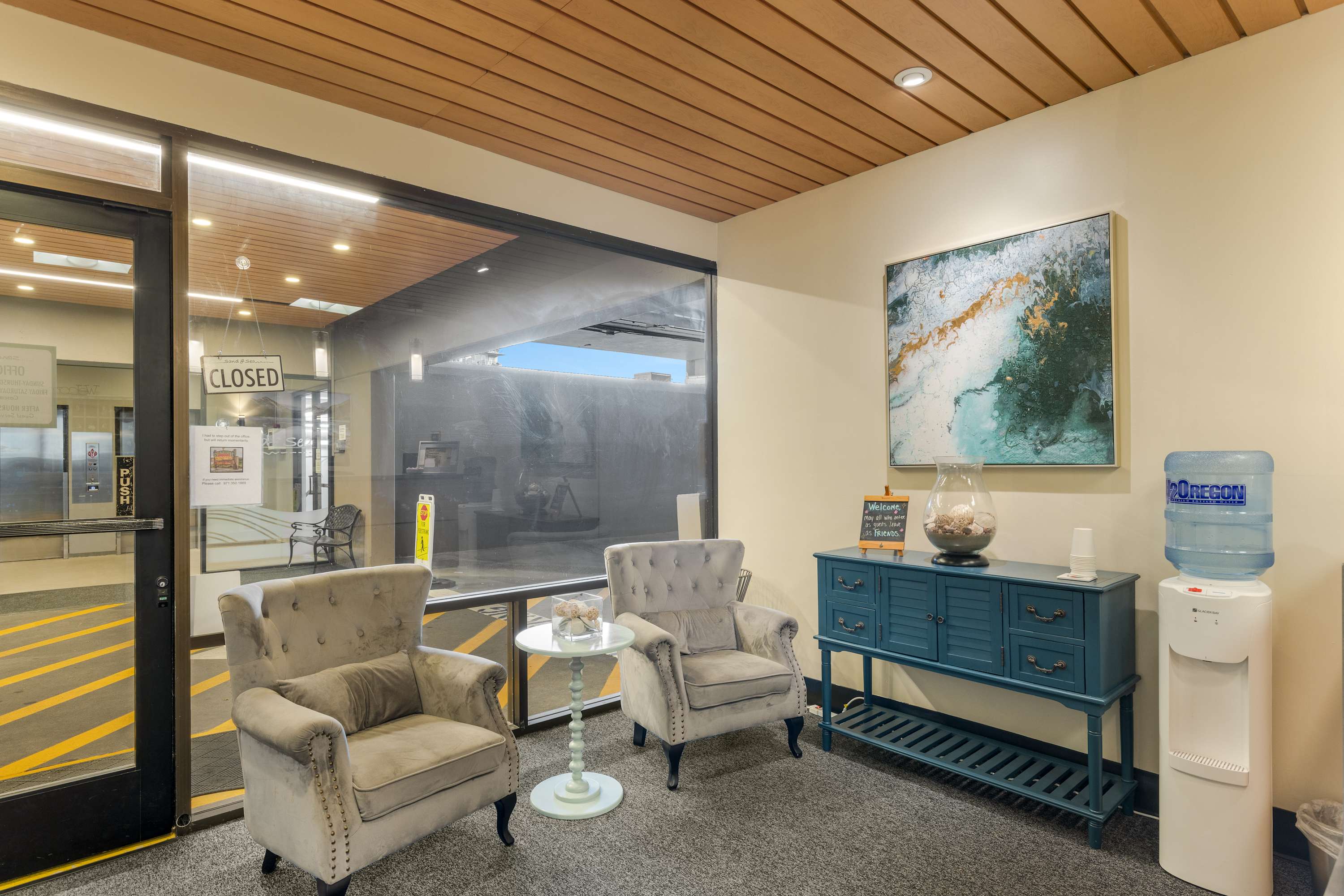 Cozy seating area in the lobby with two upholstered armchairs, a small round side table, and a decorative console table with a water cooler. Large windows overlook the exterior parking area.