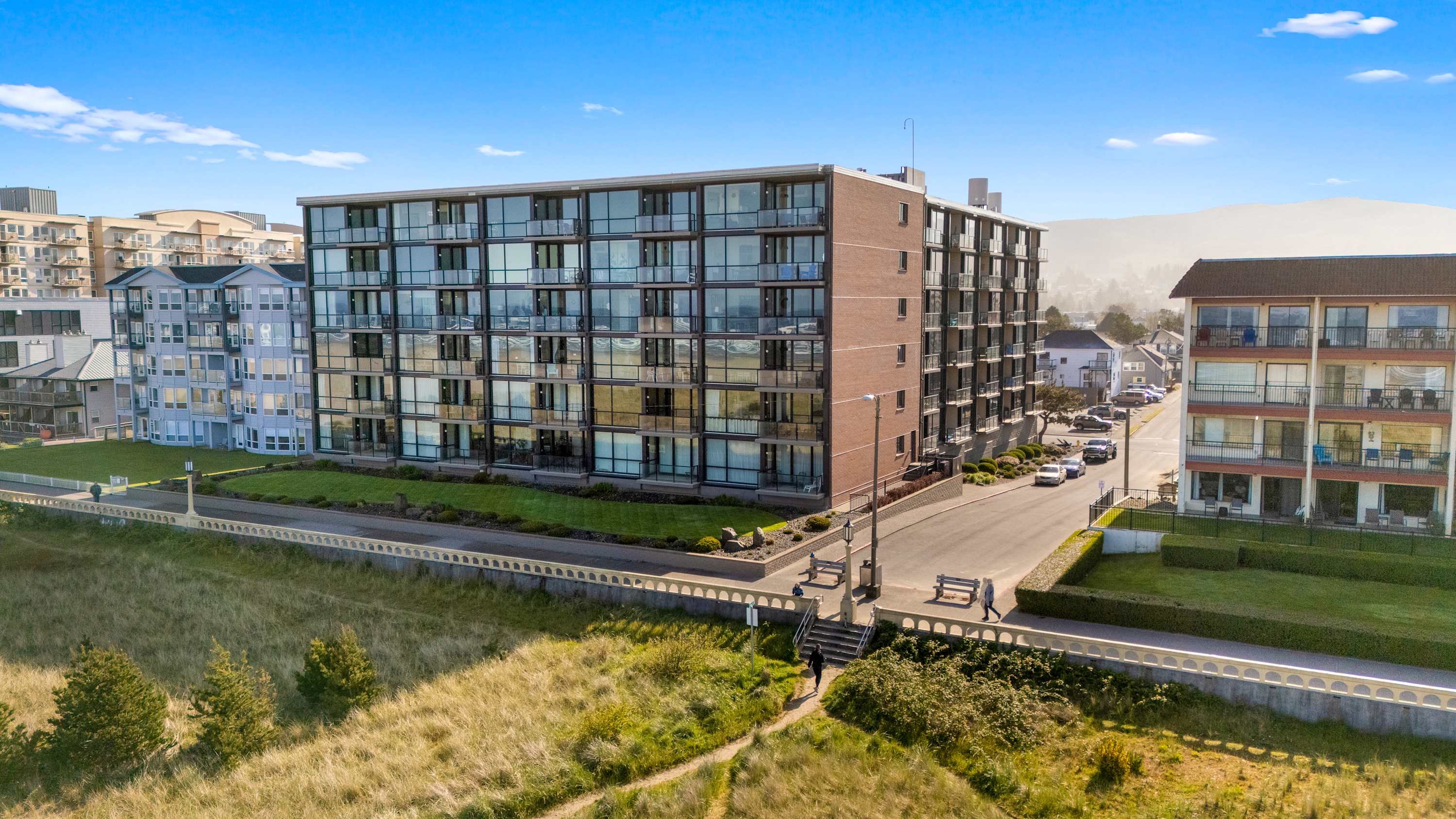 Aerial view of the condominium building from the beach side, showcasing its brick and glass architecture alongside a paved promenade and grassy dunes leading to the ocean. Mountains are visible in the distance.