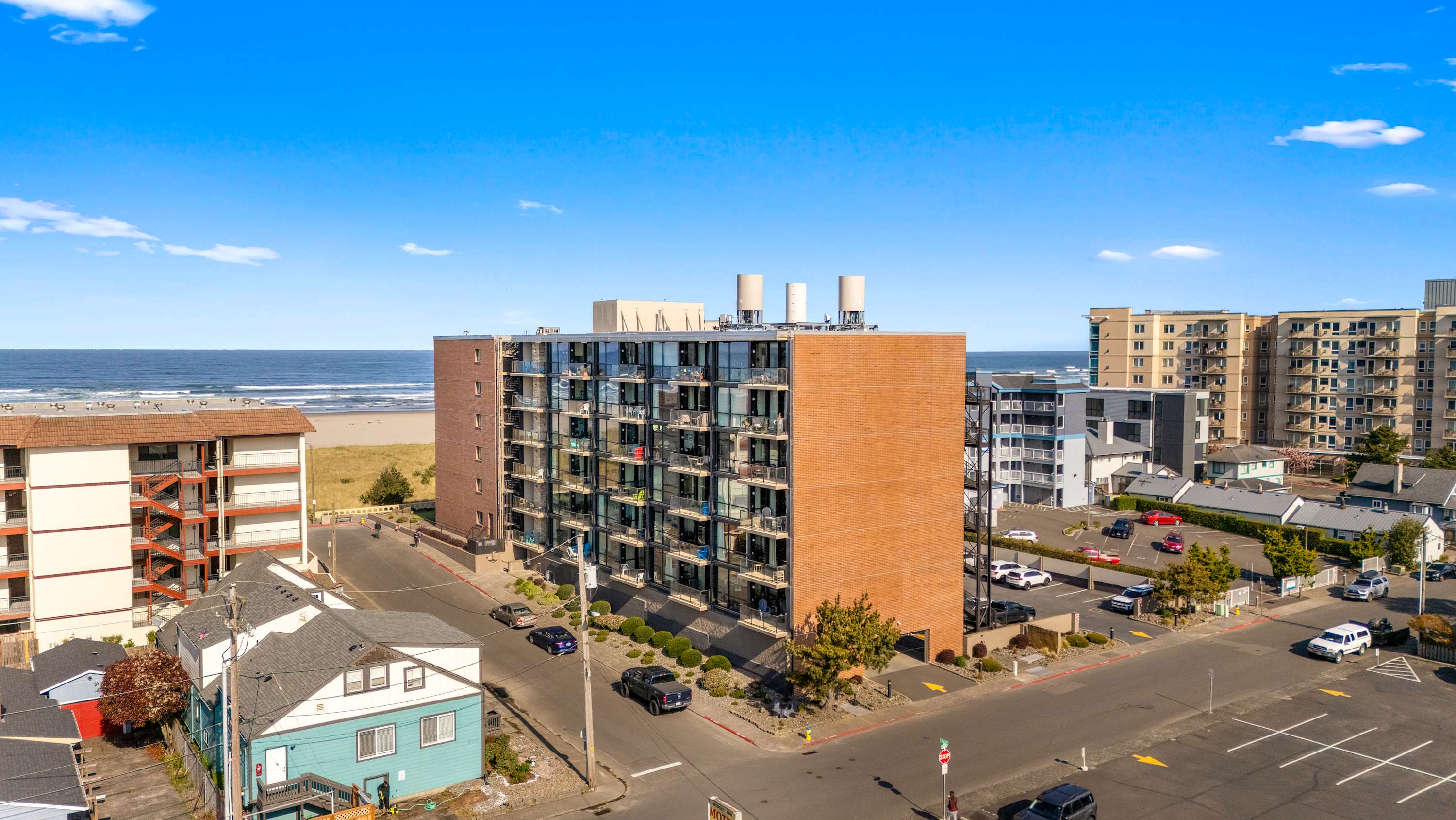 Aerial view of a multi-story brick and glass condominium building situated directly adjacent to a sandy beach and the ocean under a clear blue sky. Surrounding streets and other buildings are visible.