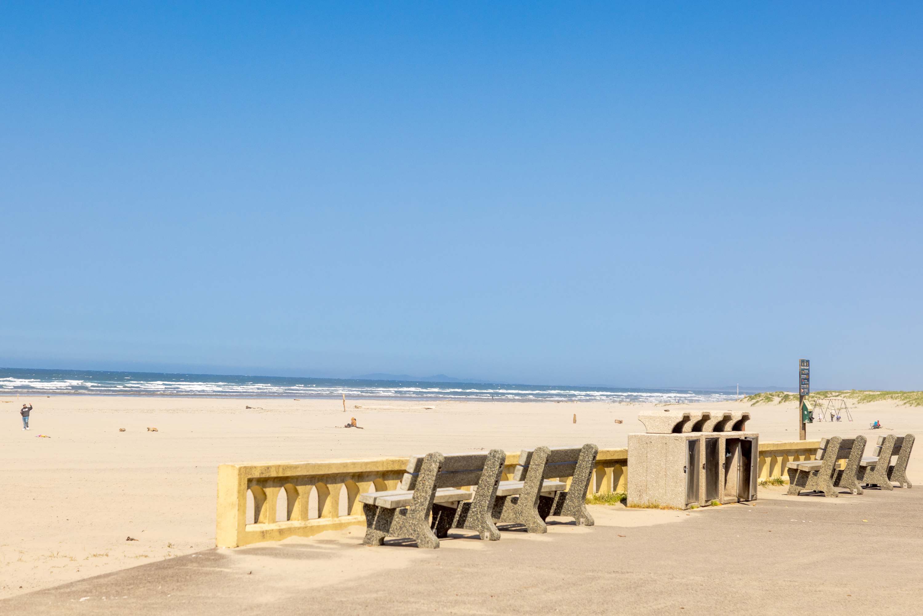 Wide shot of a vast sandy beach with gentle ocean waves under a clear blue sky, featuring a concrete promenade with multiple benches and trash receptacles. People are visible in the distance enjoying the beach.