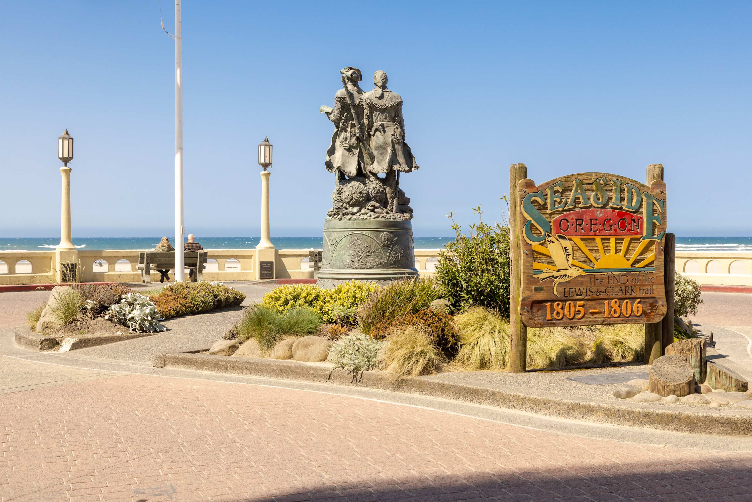 Iconic "Seaside Oregon" welcome sign and the Lewis & Clark monument on a paved plaza overlooking the ocean. Vintage-style streetlights and lush landscaping frame the historical scene.