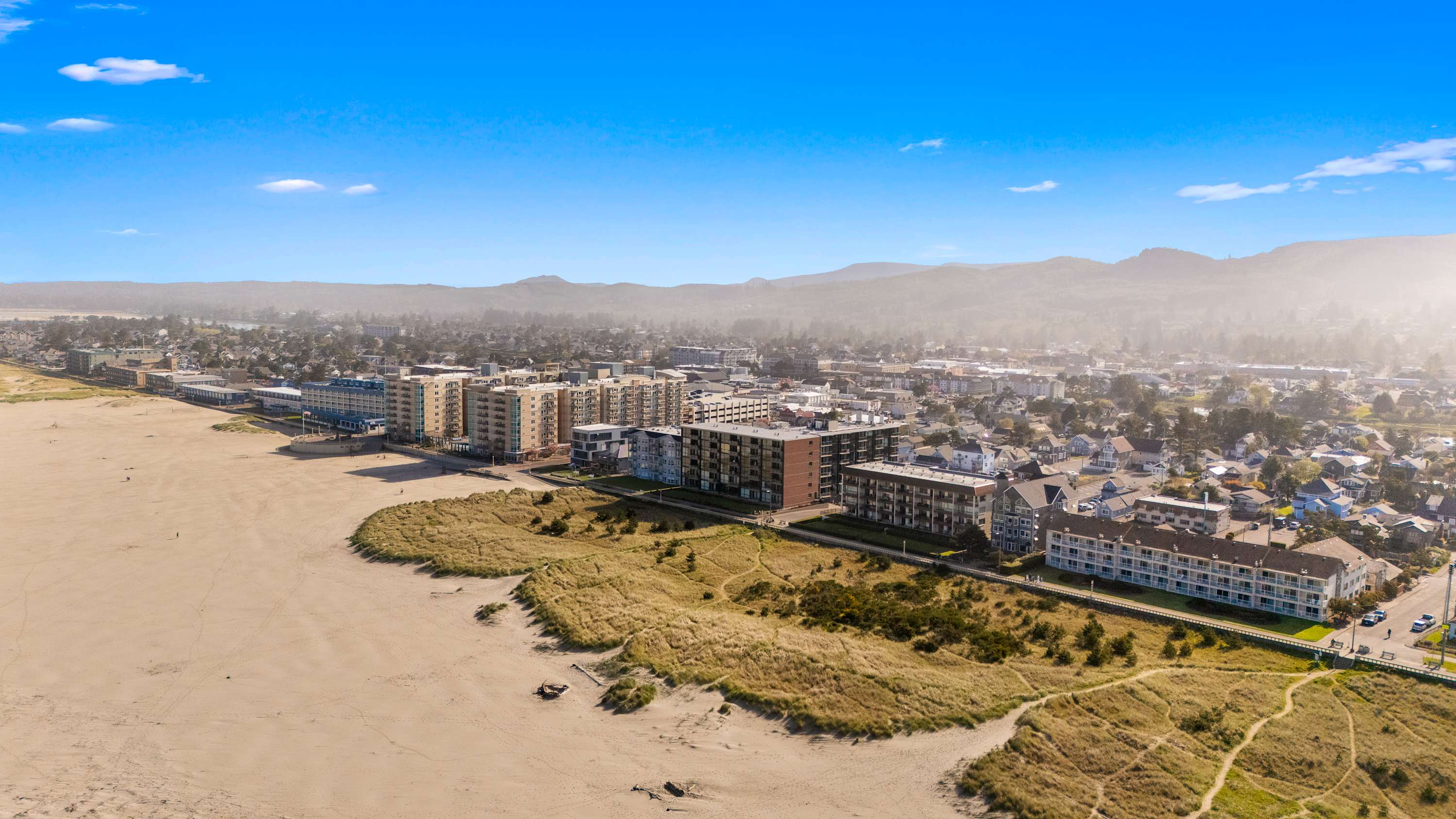 Expansive aerial view of the Seaside, Oregon coastline, featuring a long sandy beach, coastal dunes, and numerous buildings stretching along the shore and into the town, with mountains in the background.