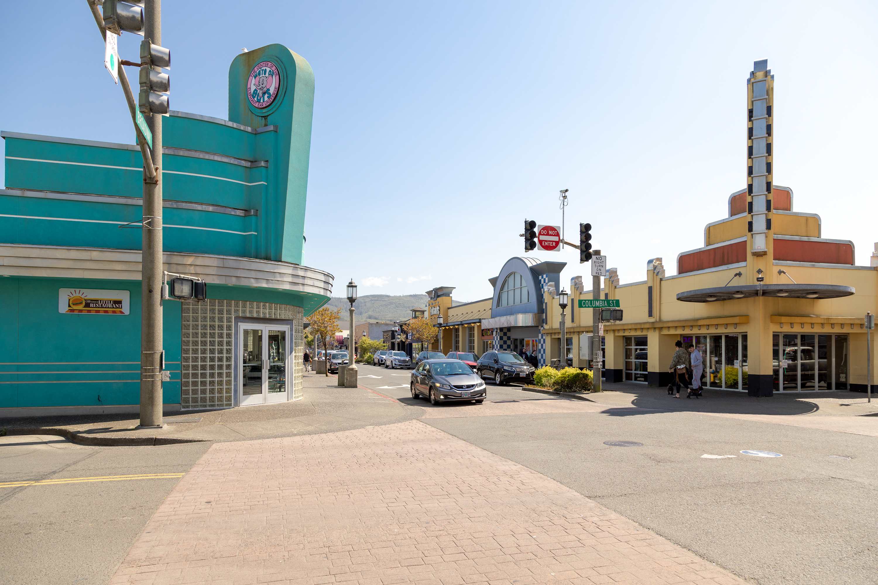 Street view of a vibrant commercial area in Seaside, Oregon, featuring retro-style buildings with unique architecture and signage, cars on the street, and pedestrians enjoying the sunny day.