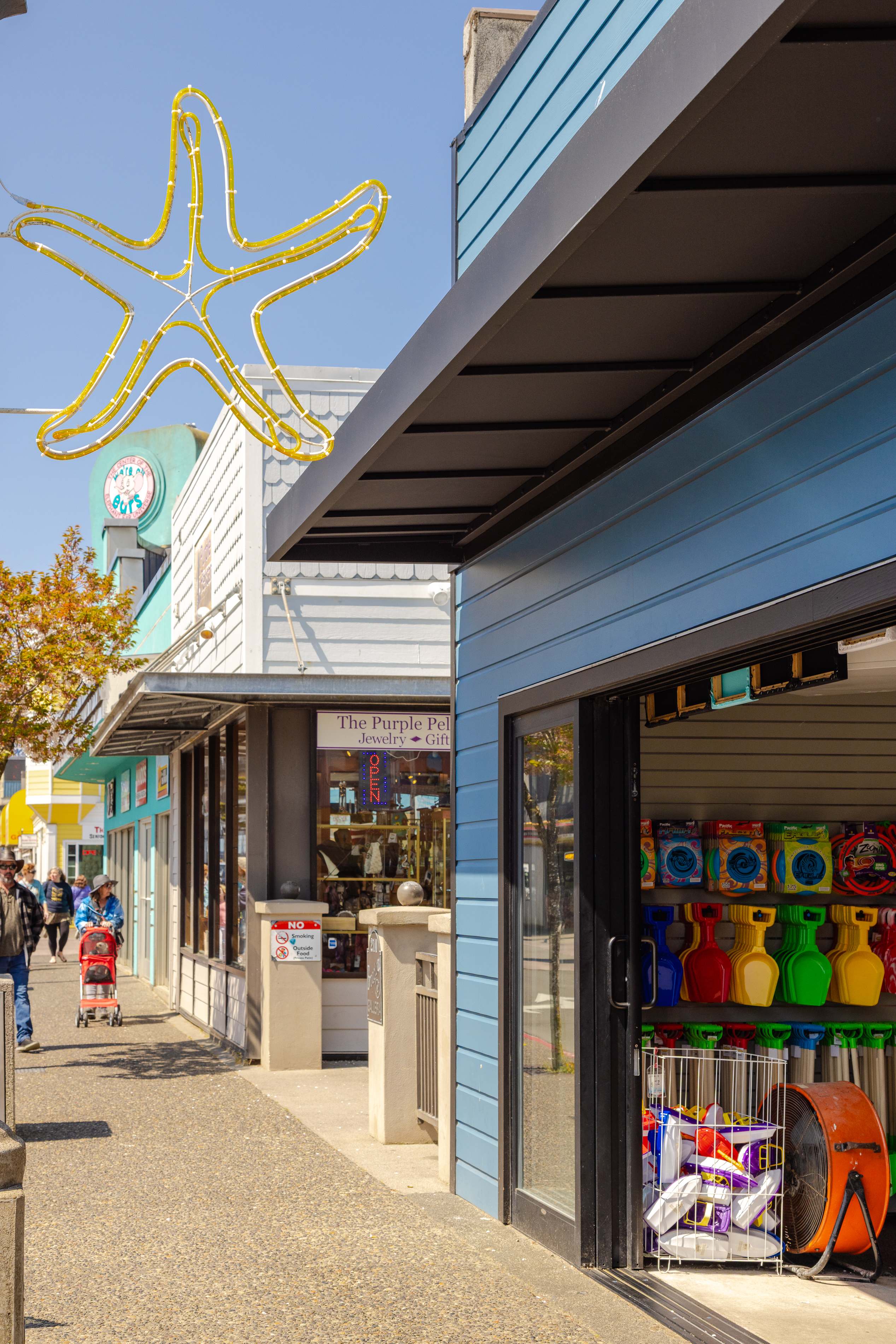 A vibrant street scene features a large yellow neon starfish sign overhead, with various storefronts including a blue-sided building displaying beach toys and an open jewelry shop. Pedestrians stroll along the sunny sidewalk.