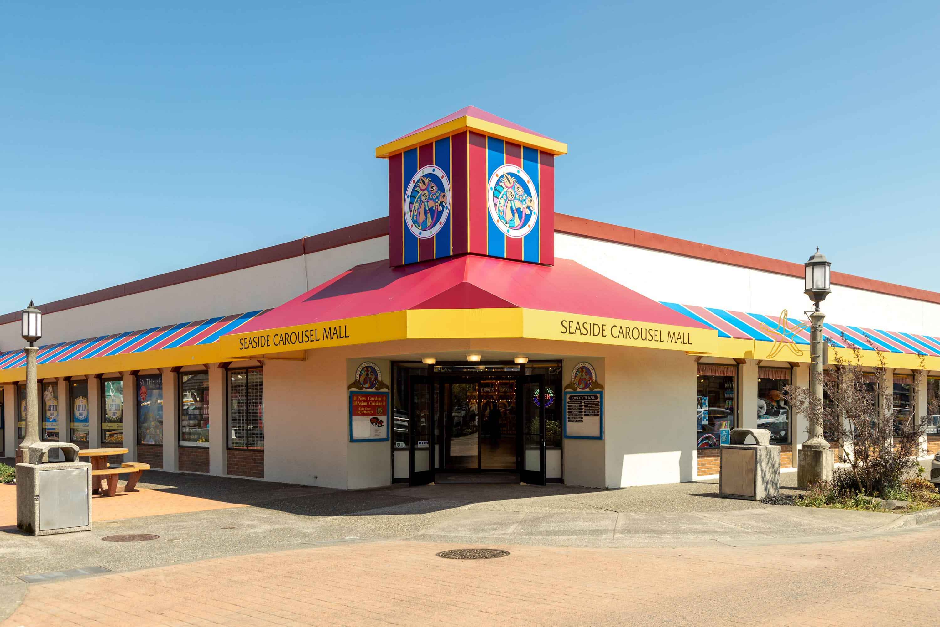 The exterior of the "Seaside Carousel Mall" features a vibrant red and yellow roof with blue and white striped awnings, and a prominent colorful tower above the main entrance. The building is set against a clear blue sky.