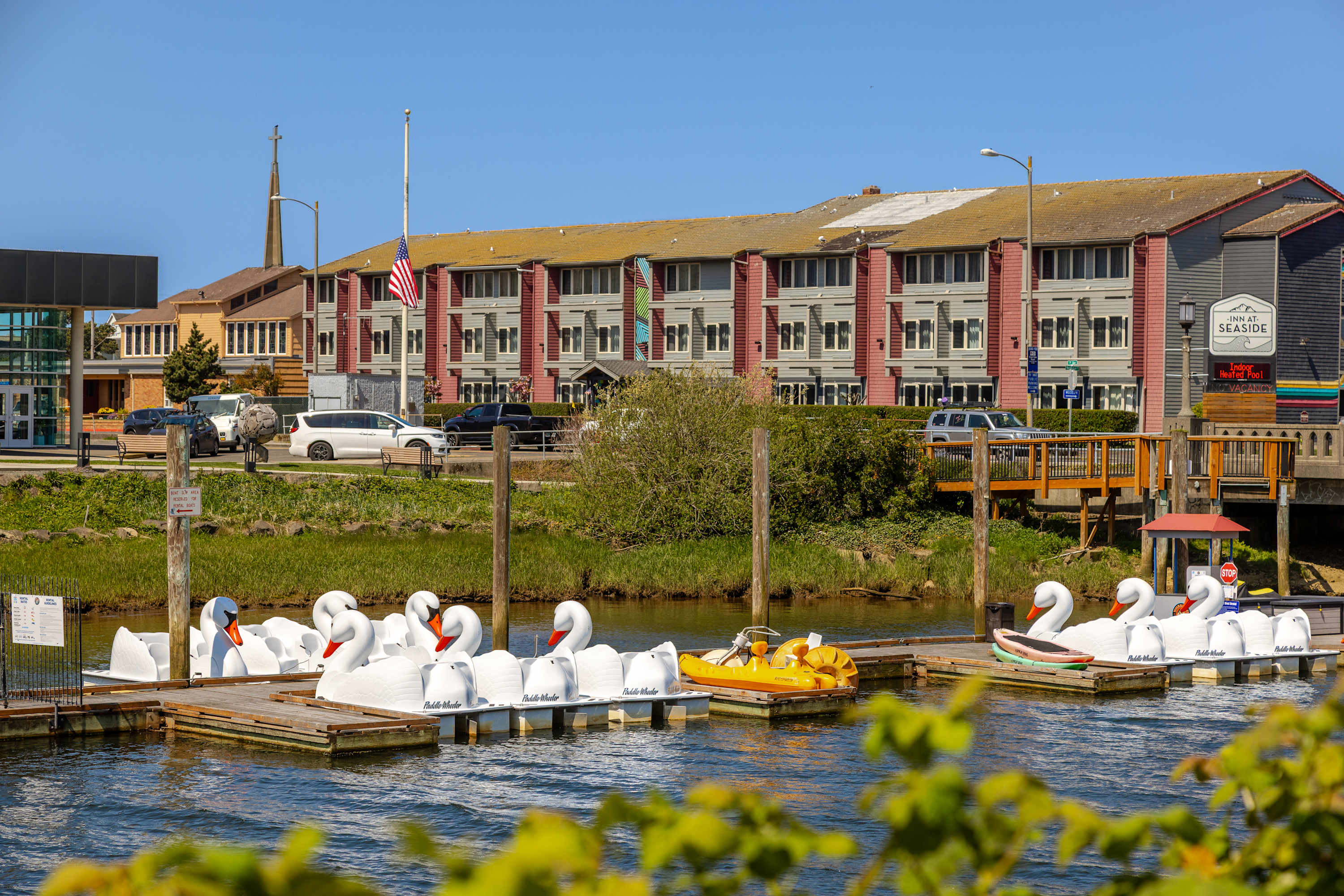 A sunny dock is lined with several white swan-shaped paddle boats and a yellow paddle boat, floating on calm water. In the background, multi-story buildings and lush greenery line the shore under a clear blue sky.