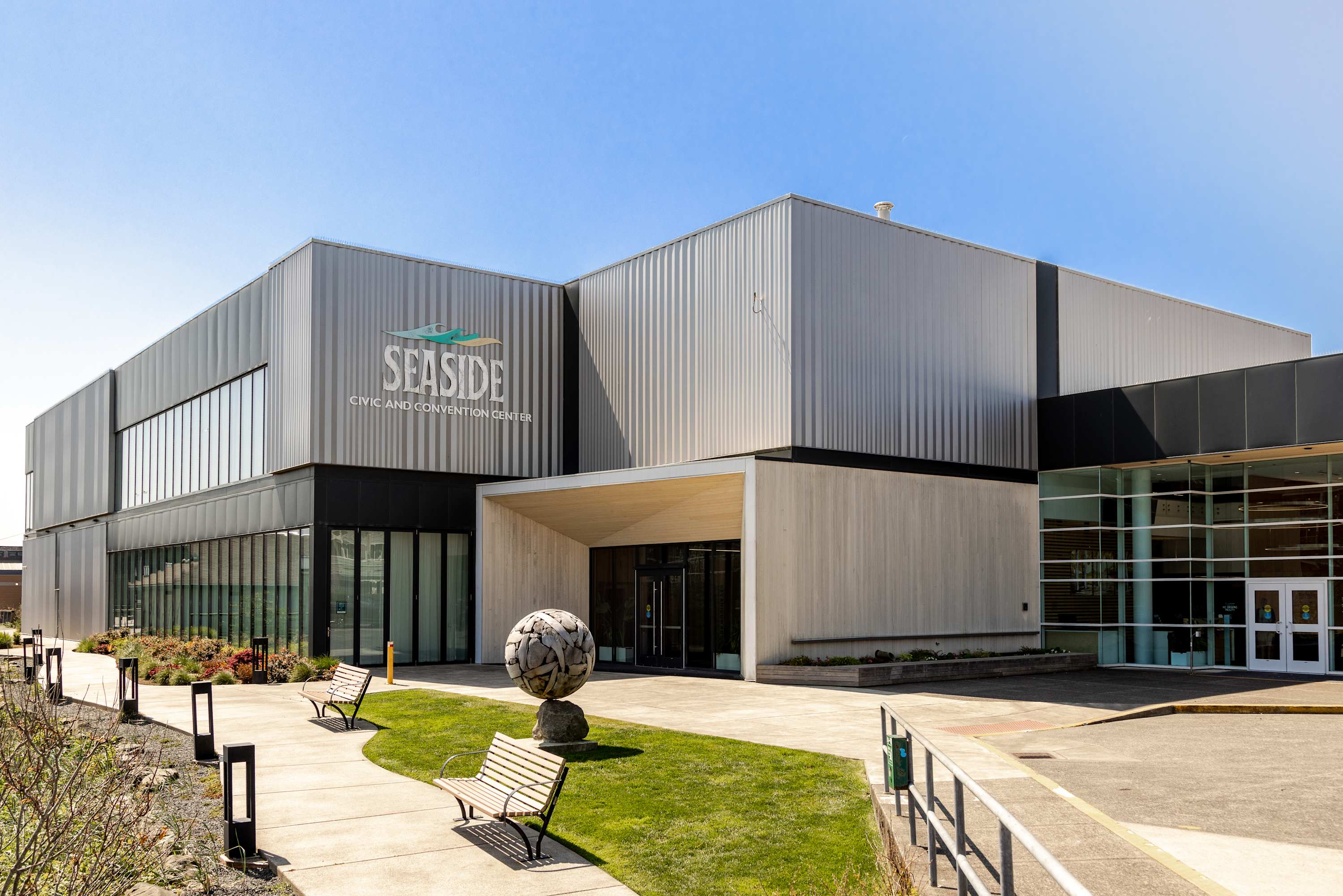 The modern "Seaside Civic and Convention Center" building features a striking silver corrugated facade and large glass windows, with a welcoming entrance area. Benches and landscaped greenery adorn the paved walkway leading to the building.