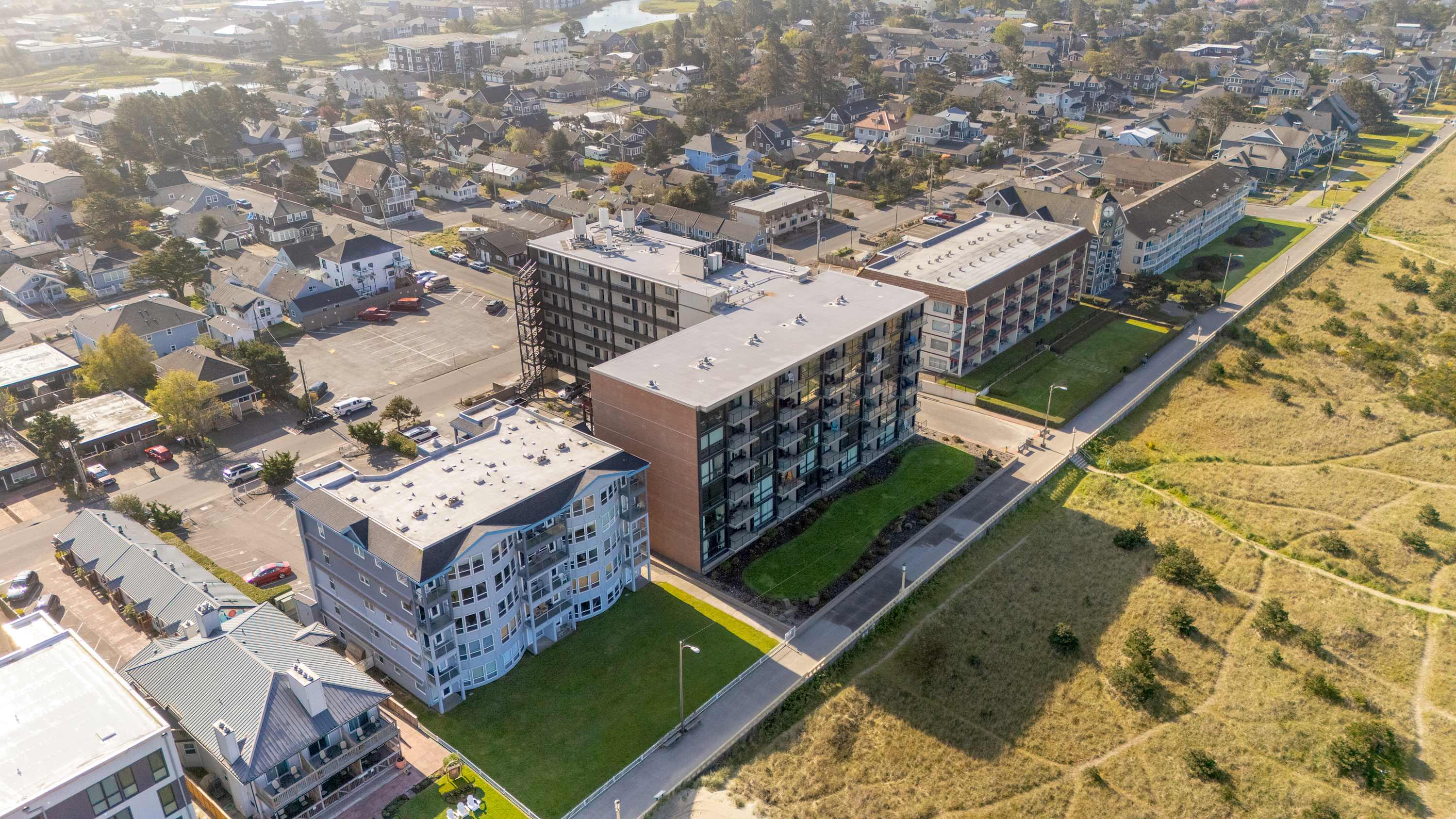 An aerial view captures several large condominium buildings with modern architecture, nestled within a coastal town alongside residential homes. A wide expanse of green grass and sandy dunes separates the buildings from the distant ocean.