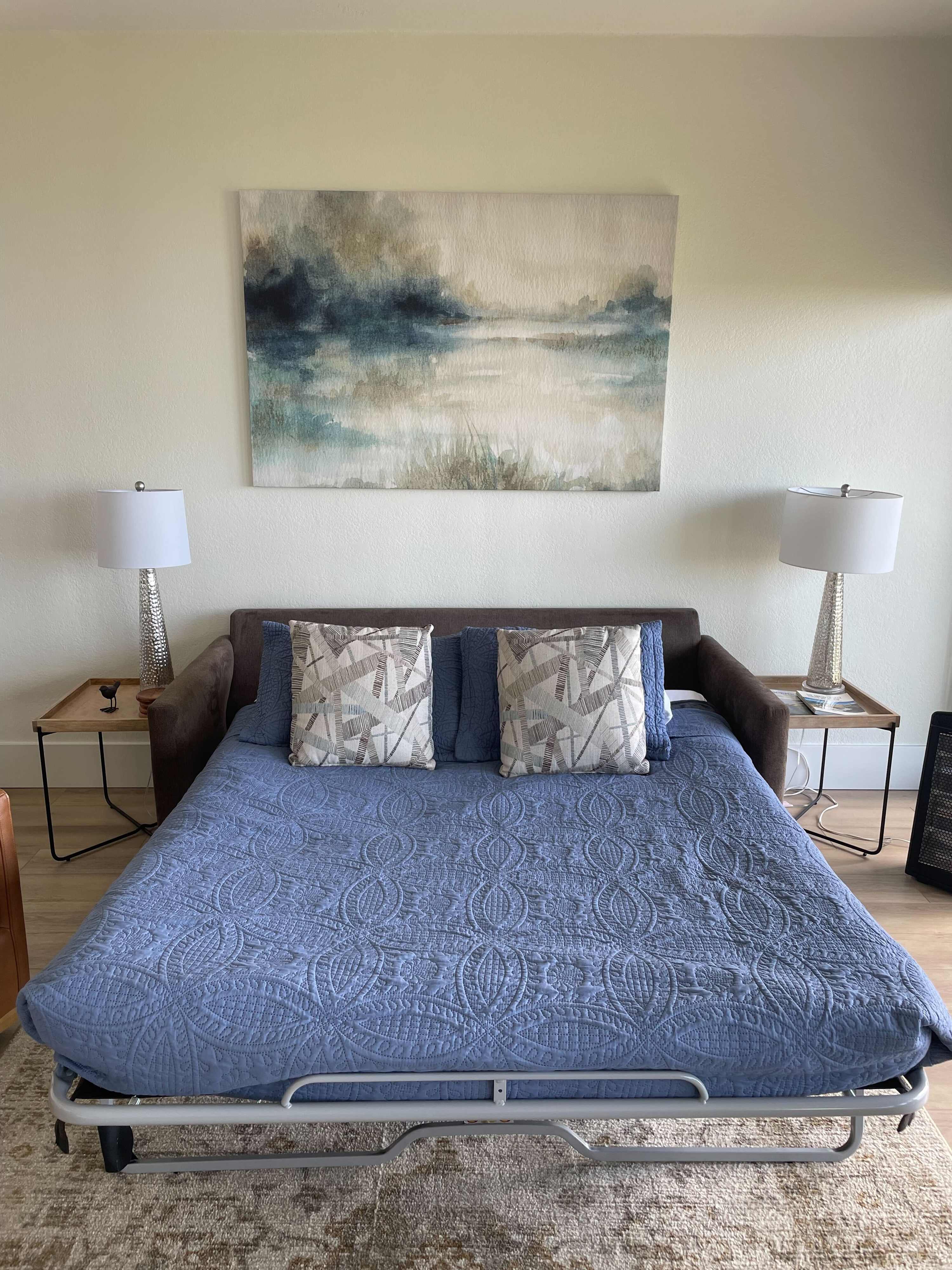 Living room transformed into a sleeping area with a sofa bed pulled out and made with a blue quilted cover, flanked by two side tables with lamps, and an abstract painting on the wall.