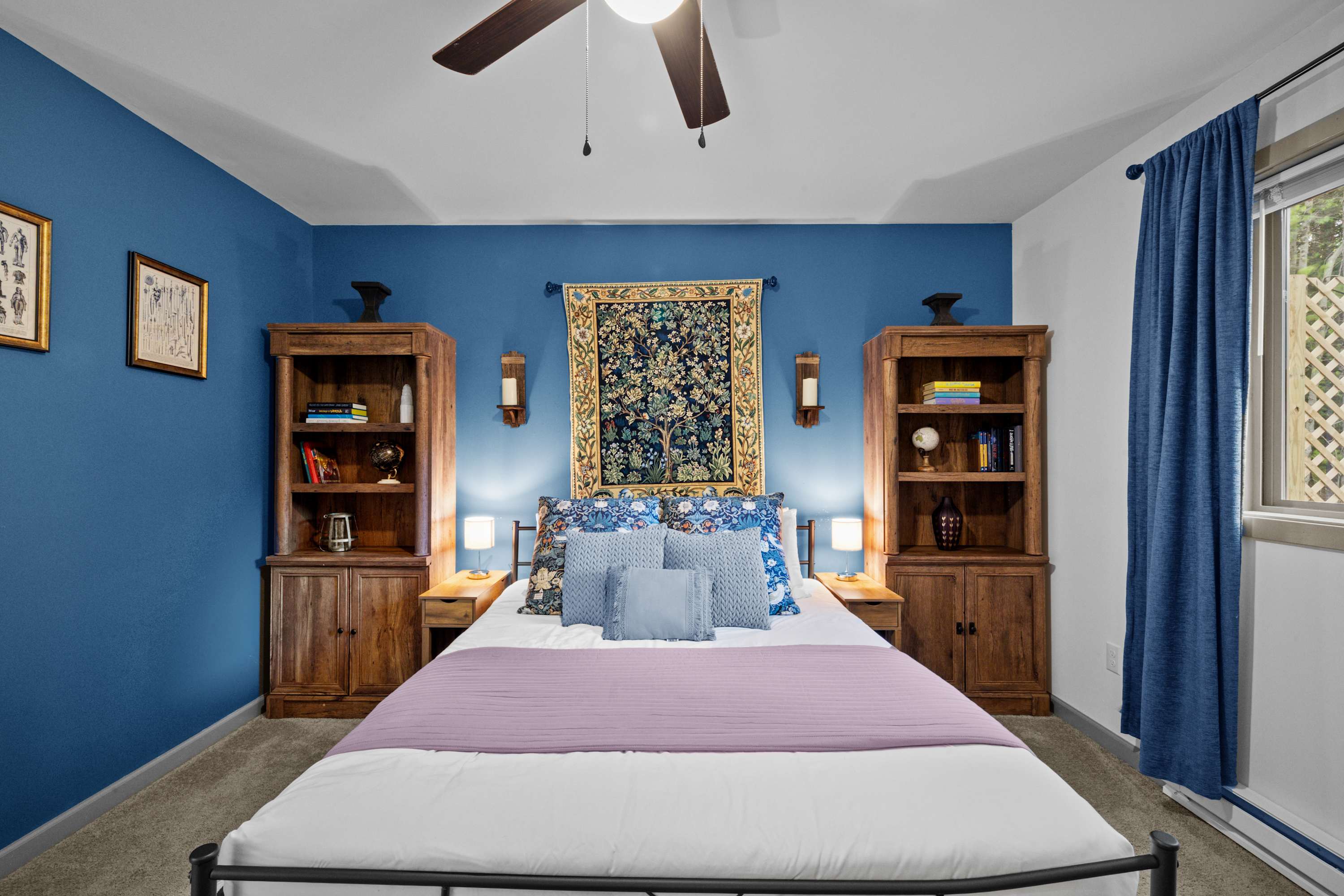 Bedroom #1 features a queen bed, soft lighting, and a stunning tapestry. The warm wooden furniture and inviting ambiance make it the perfect space to sleep and dream.