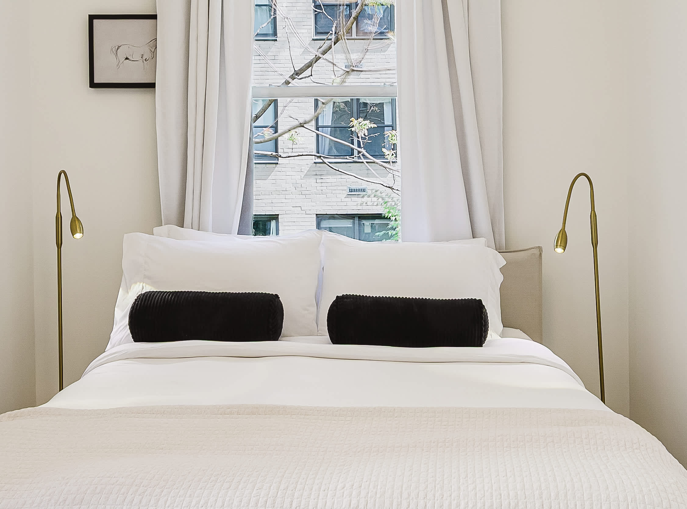 Bedroom with a white bed, black accent pillows, brass reading lamps, and a window view of a brick facade.
