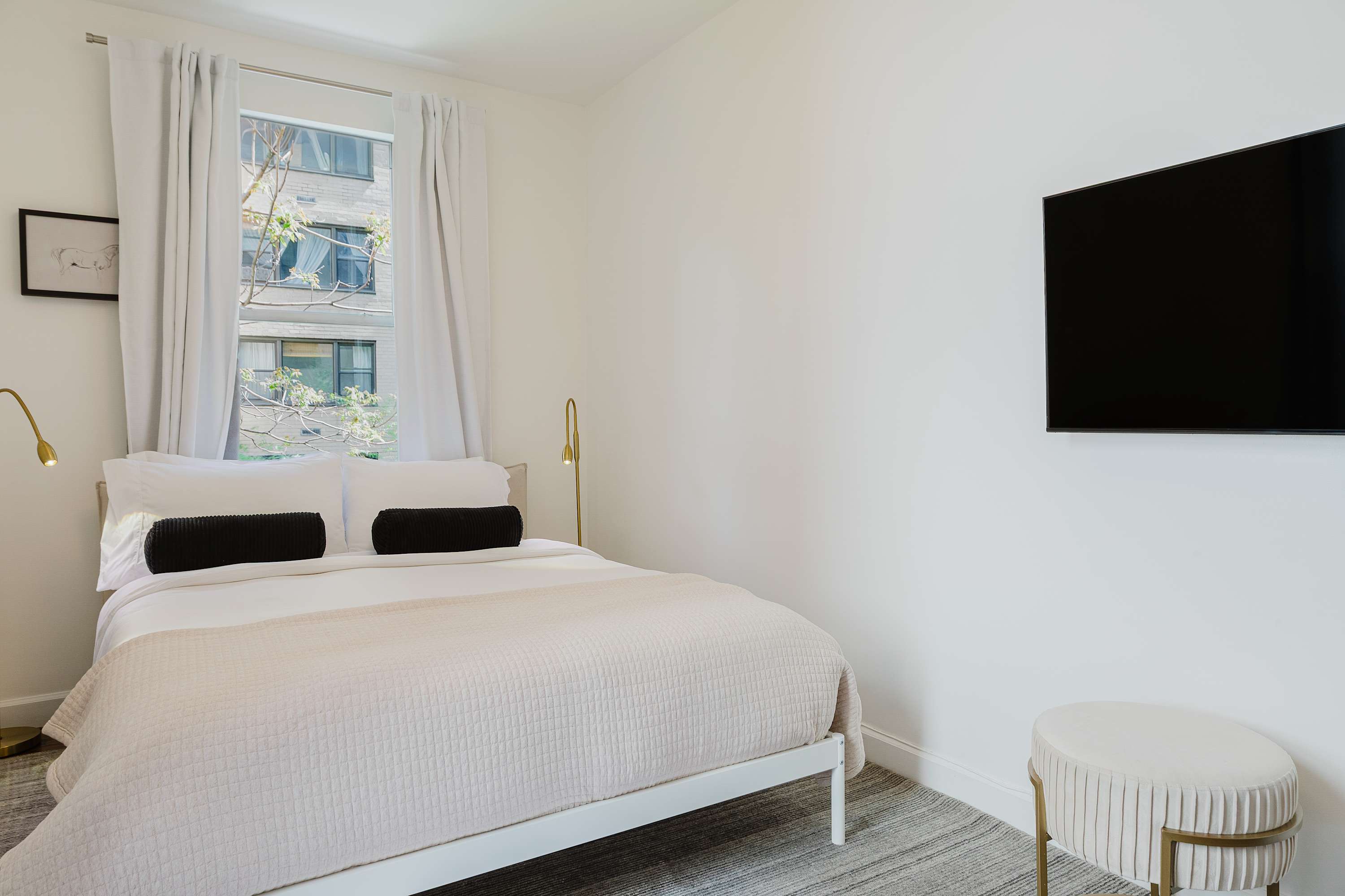 Bedroom with a white metal bed frame, wall-mounted TV, and a round tufted stool.