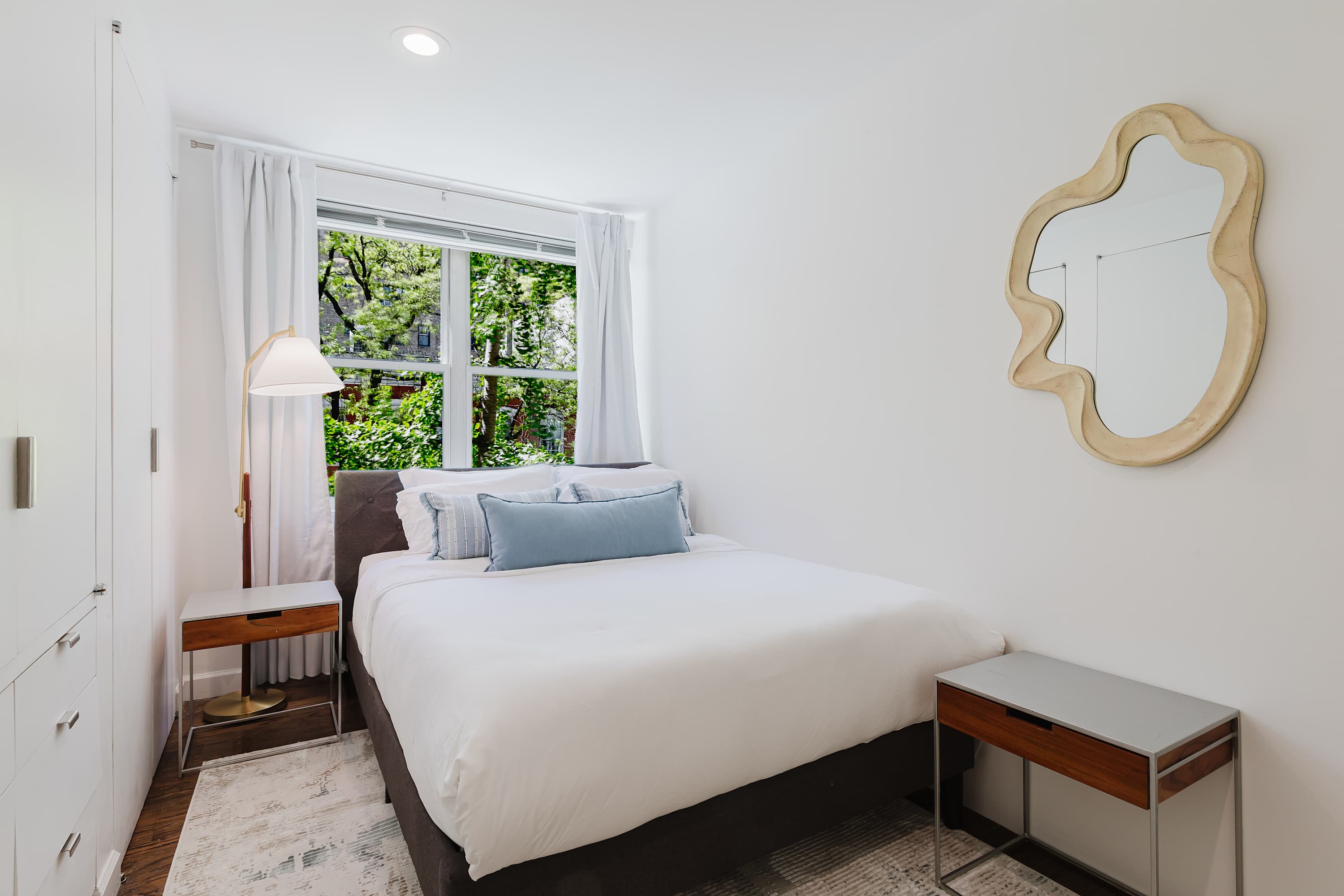 Bedroom with a white bed, a blue accent pillow, decorative mirror, and window seating.