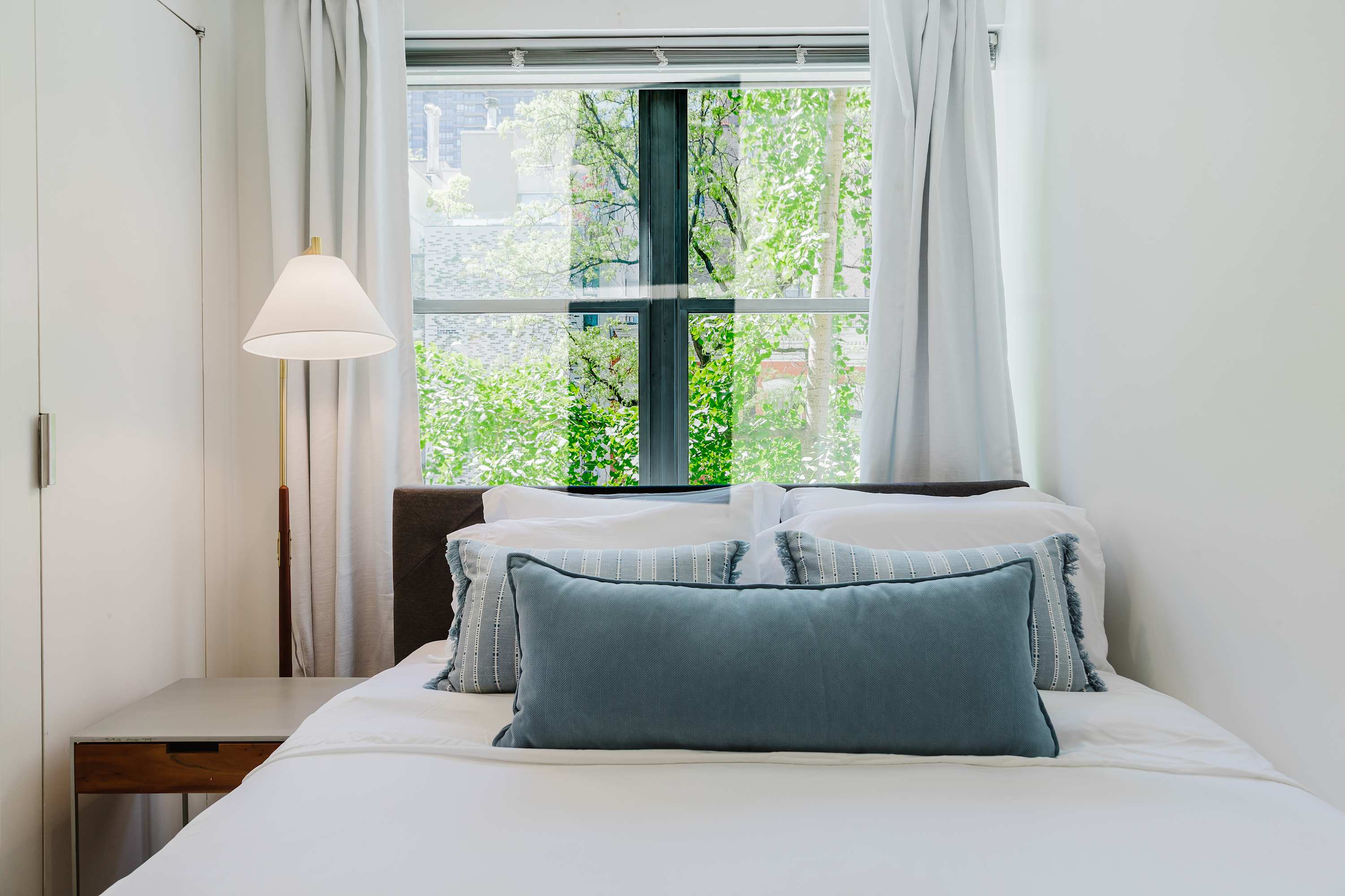 Bedroom with a white bed, blue accent pillows, and a window overlooking trees.