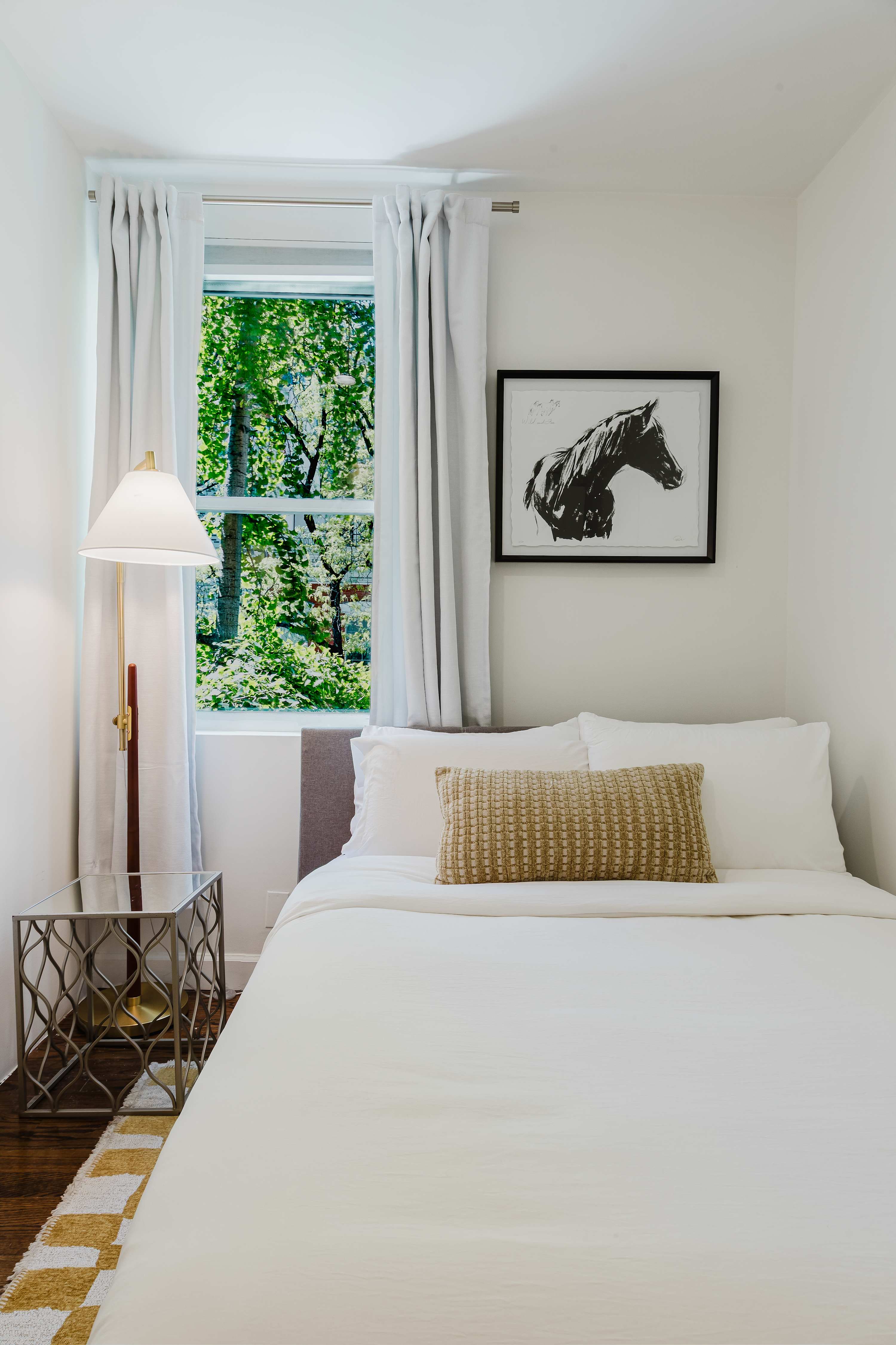 Bedroom corner with a framed horse print above the headboard and a leafy window view.