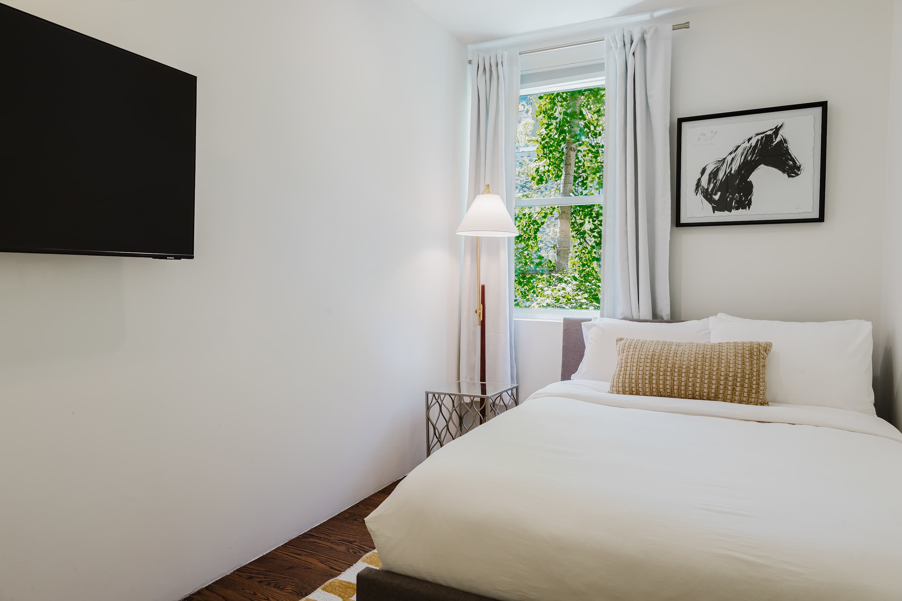 Bedroom with a white bed, framed horse print, gold reading lamp, and a window overlooking greenery.