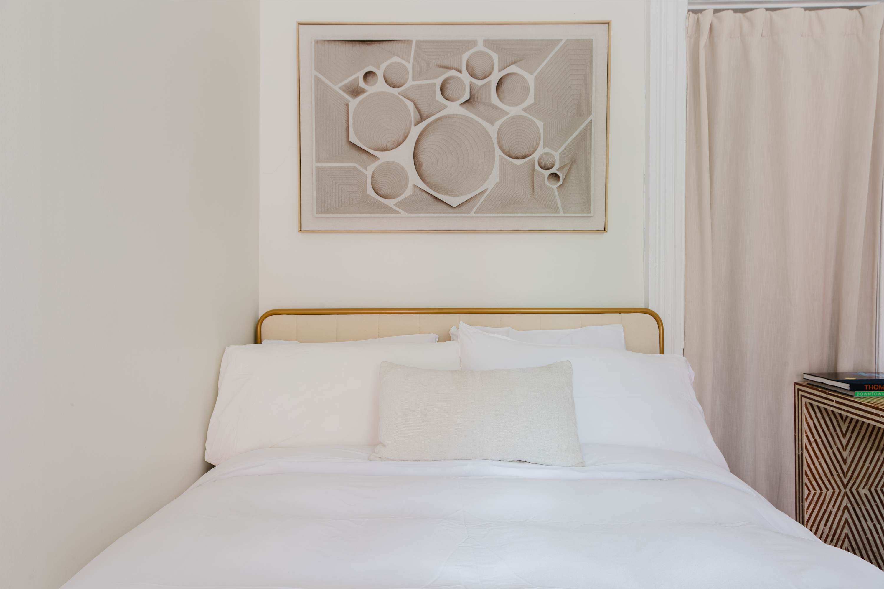 Close-up of the bed with white linens, a brass headboard, and abstract geometric wall art.