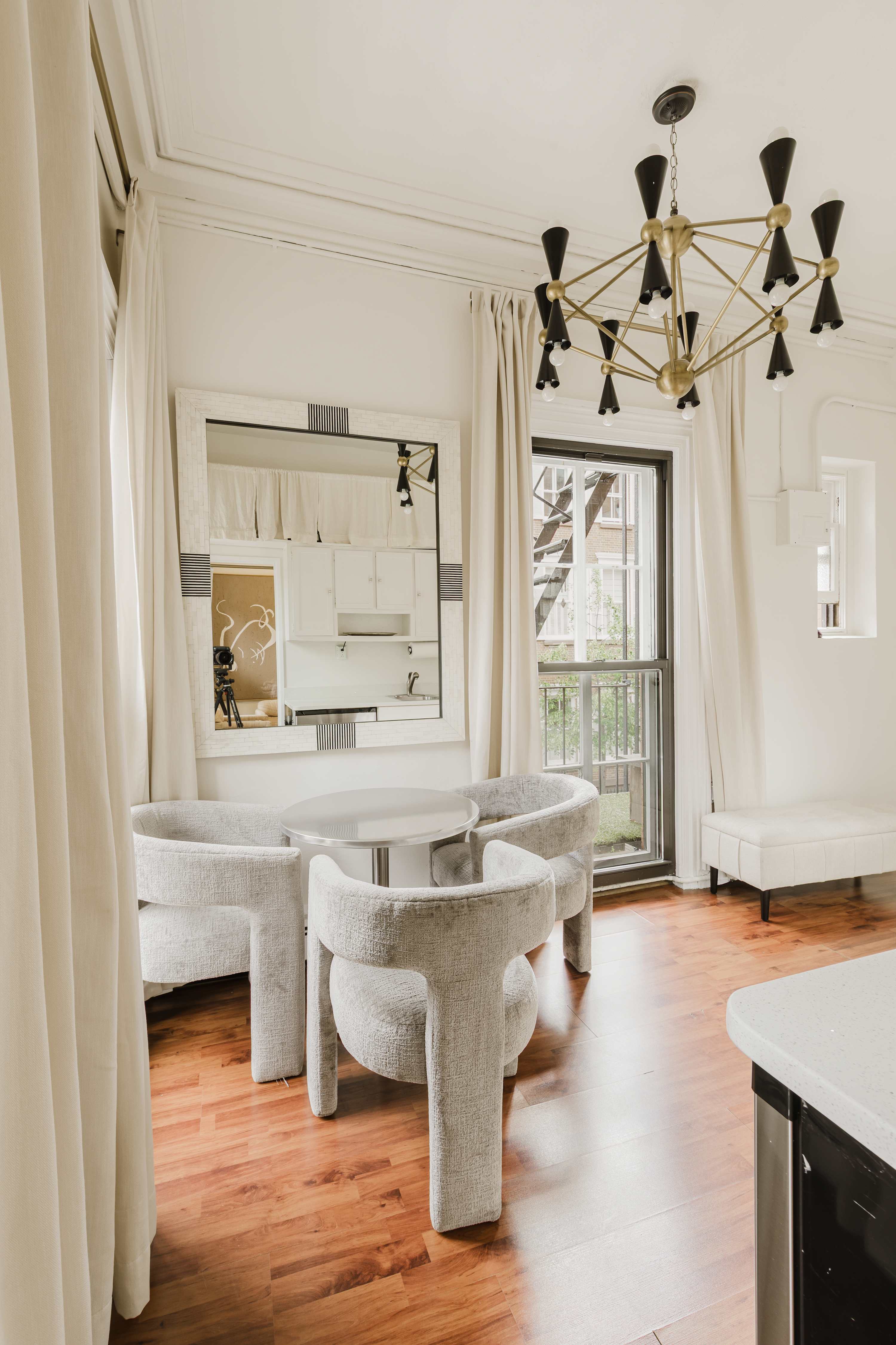 Dining area with a round marble table, curved white chairs, brass chandelier, and French doors opening to a Juliet balcony.