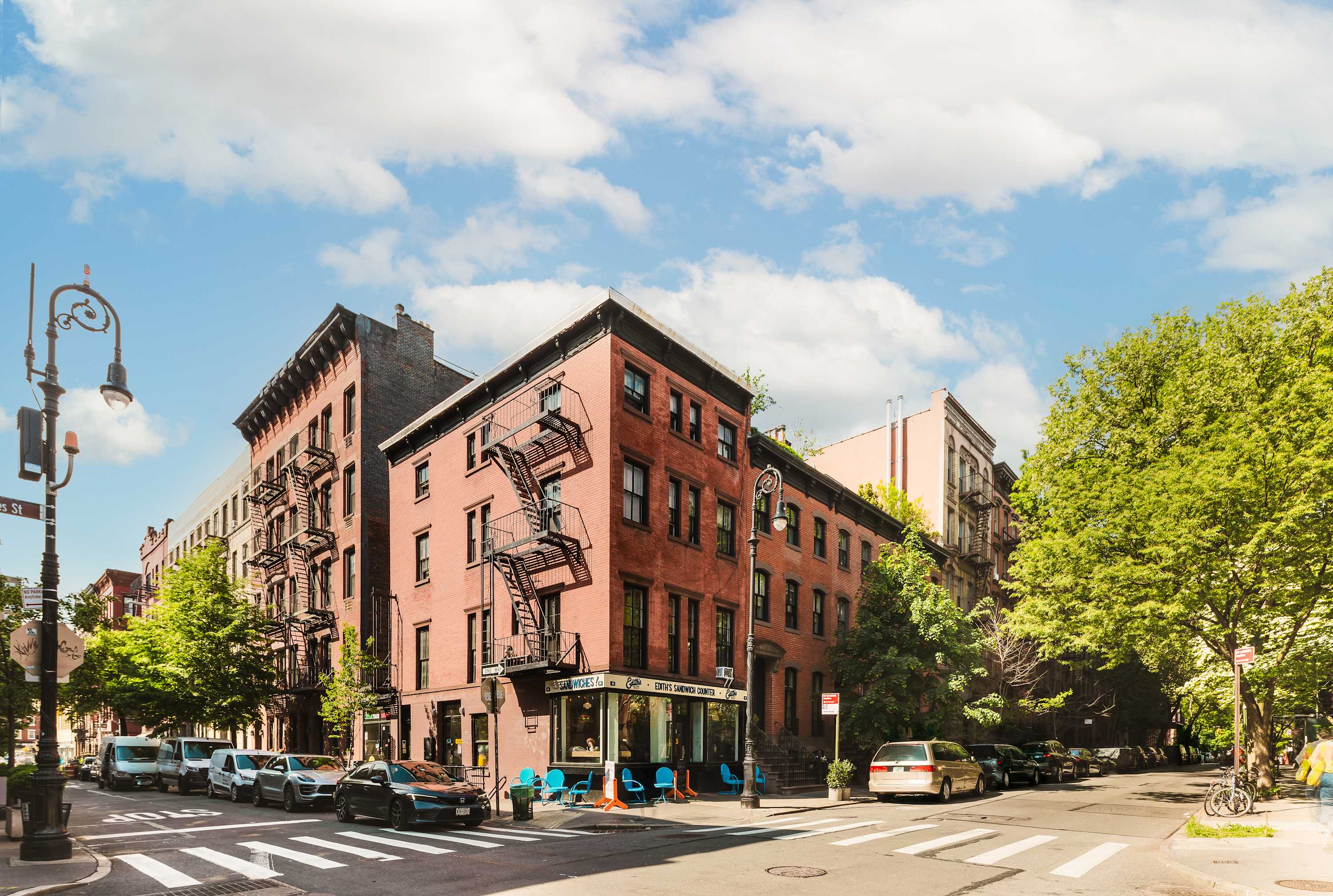 Street view of the red-brick corner building with fire escapes, a ground-floor cafe, and a crosswalk at the intersection.