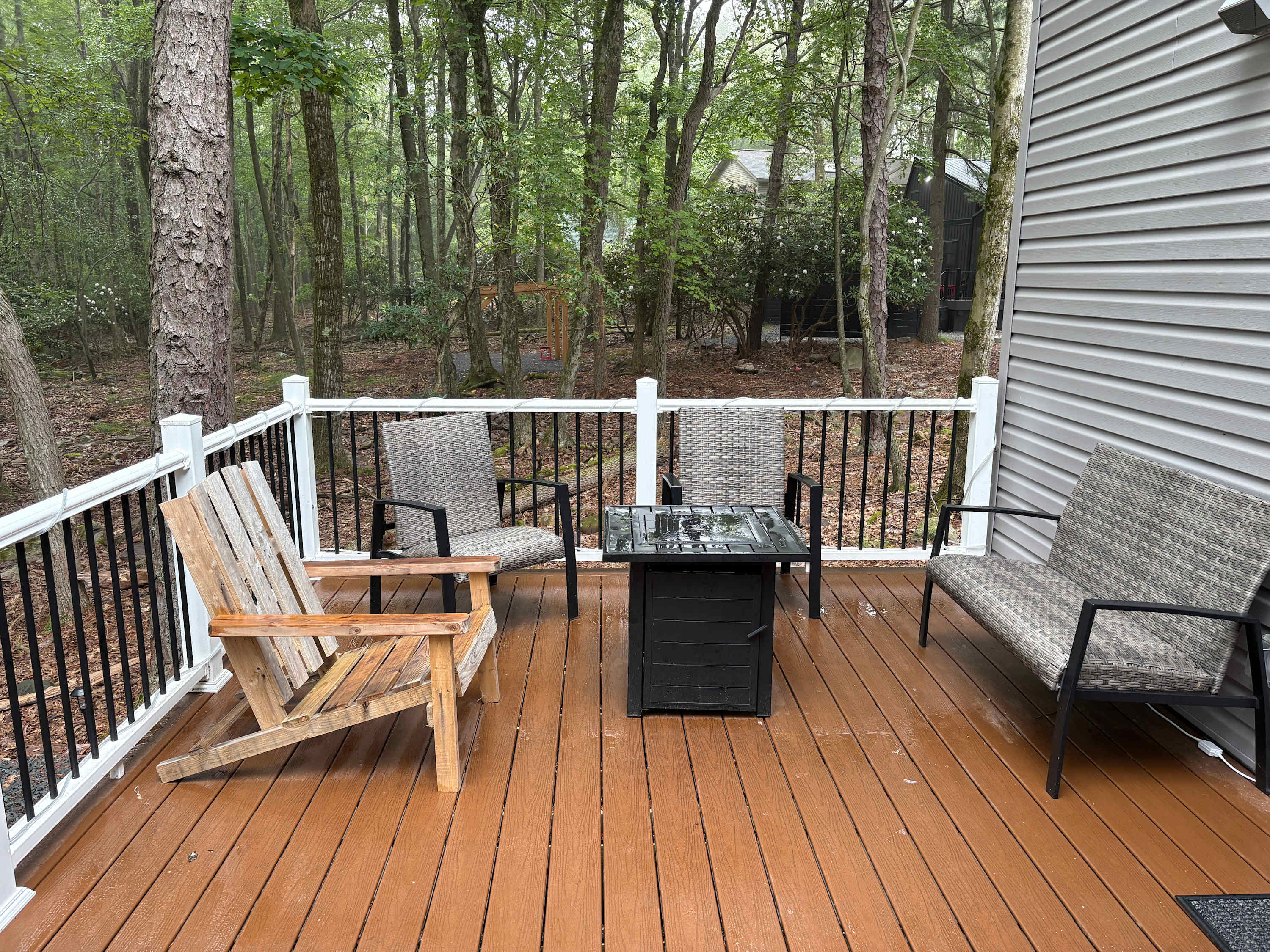 Outdoor deck seating area with fire pit table. Four outdoor chairs including wicker and wood Adirondack-style chairs. Dark stained deck with white and black railing. Wooded surroundings.