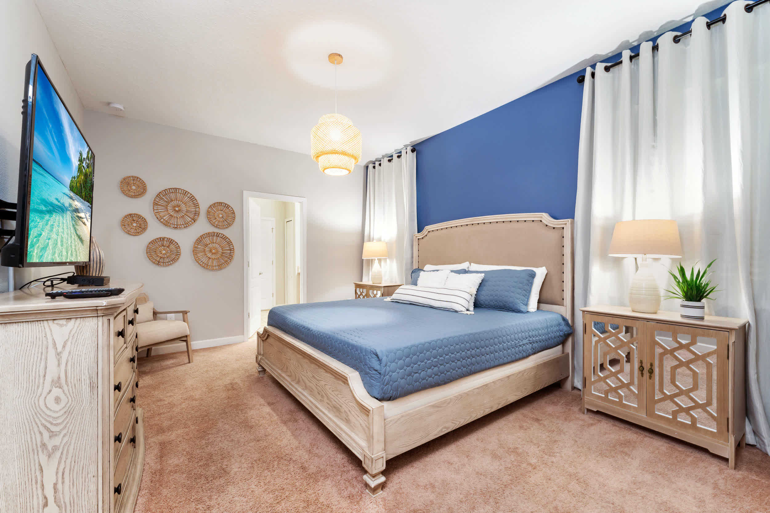 Coastal-inspired bedroom with a king-size bed, blue accent wall, tropical beach-view TV, and bright floor-to-ceiling curtains.