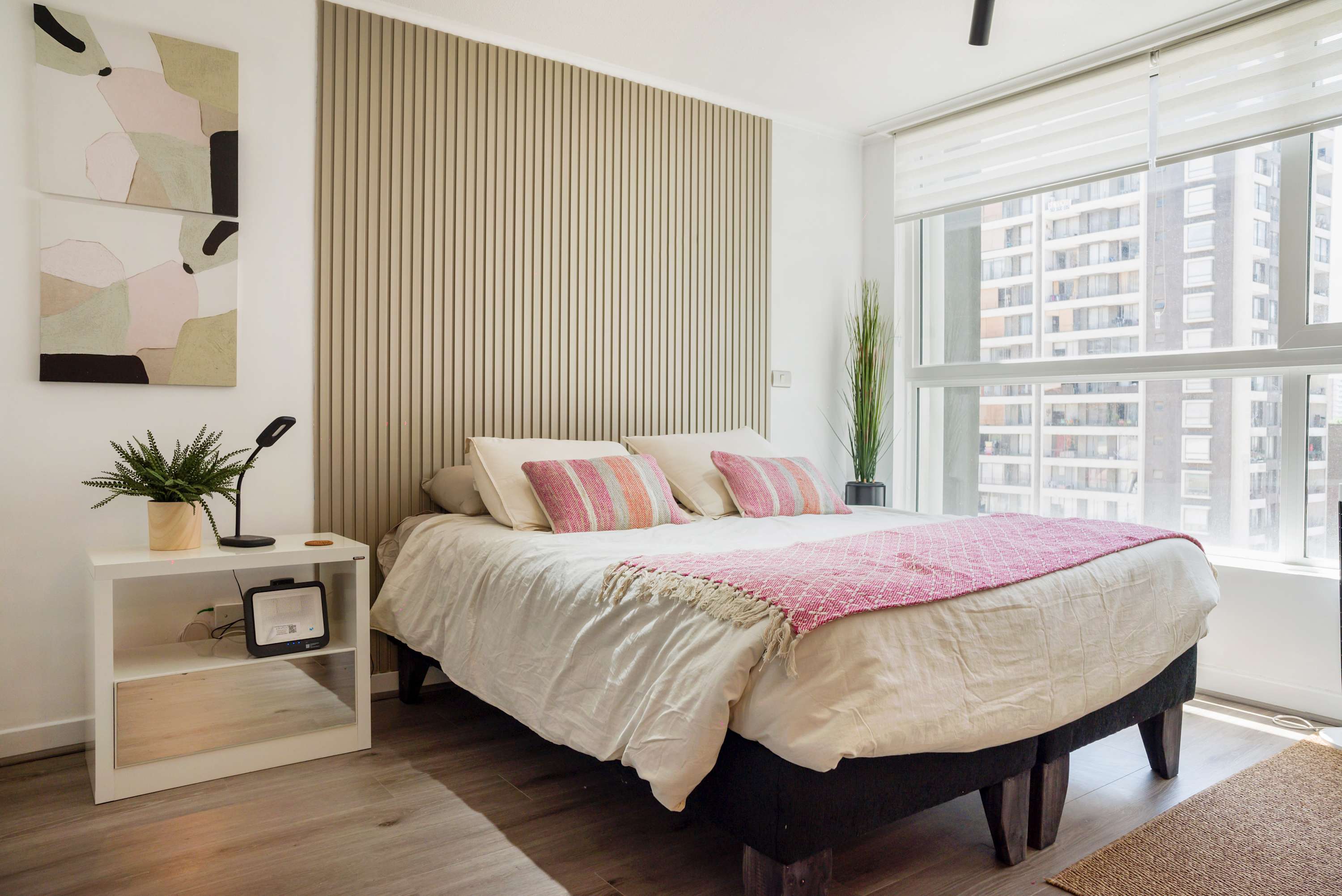 Bright studio sleeping area with a textured pink throw, layered pillows, a slatted feature wall, and floor-to-ceiling windows overlooking the city.