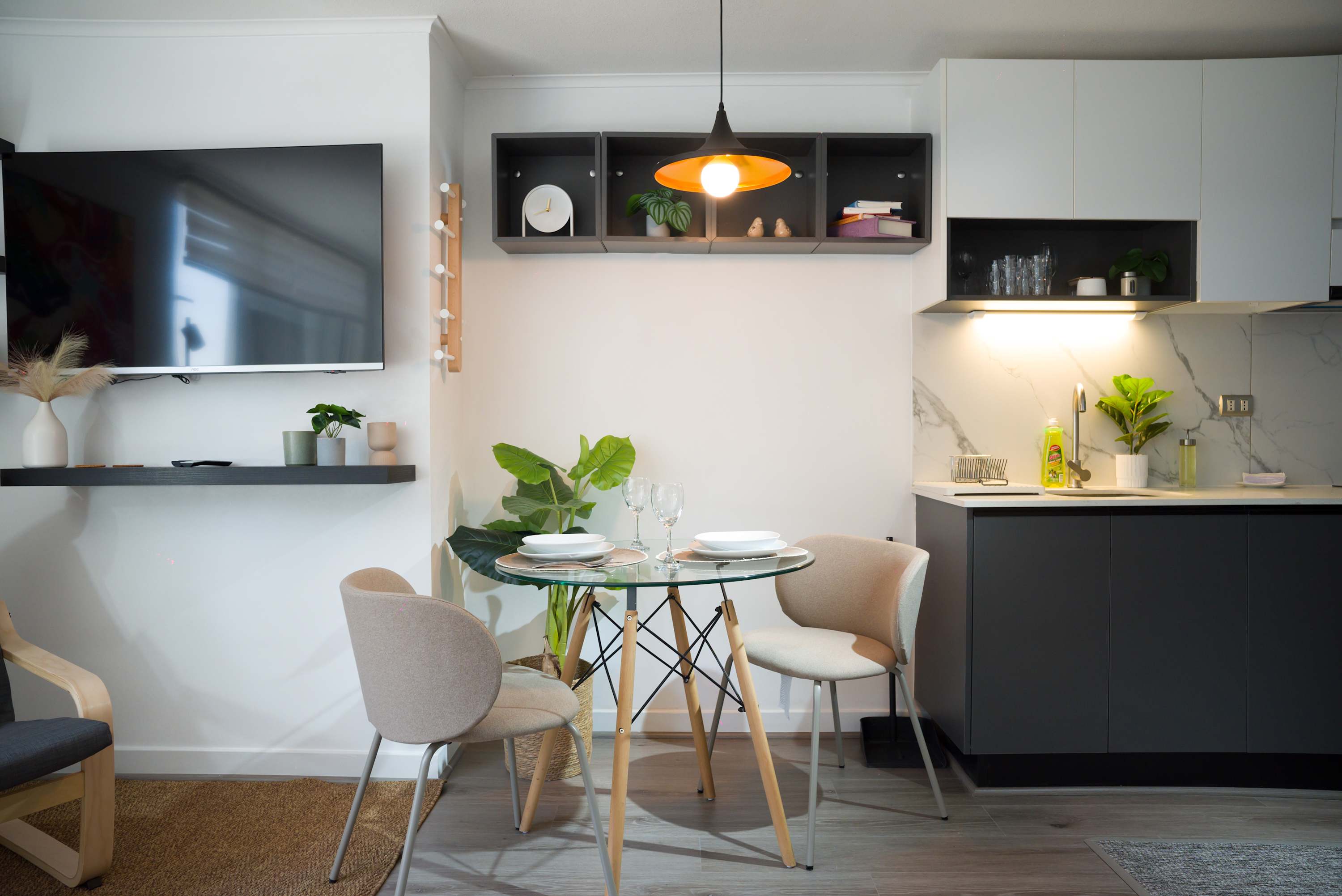 Open-plan dining area with a glass table set for two beside the dark cabinetry and marble-look kitchen backsplash.