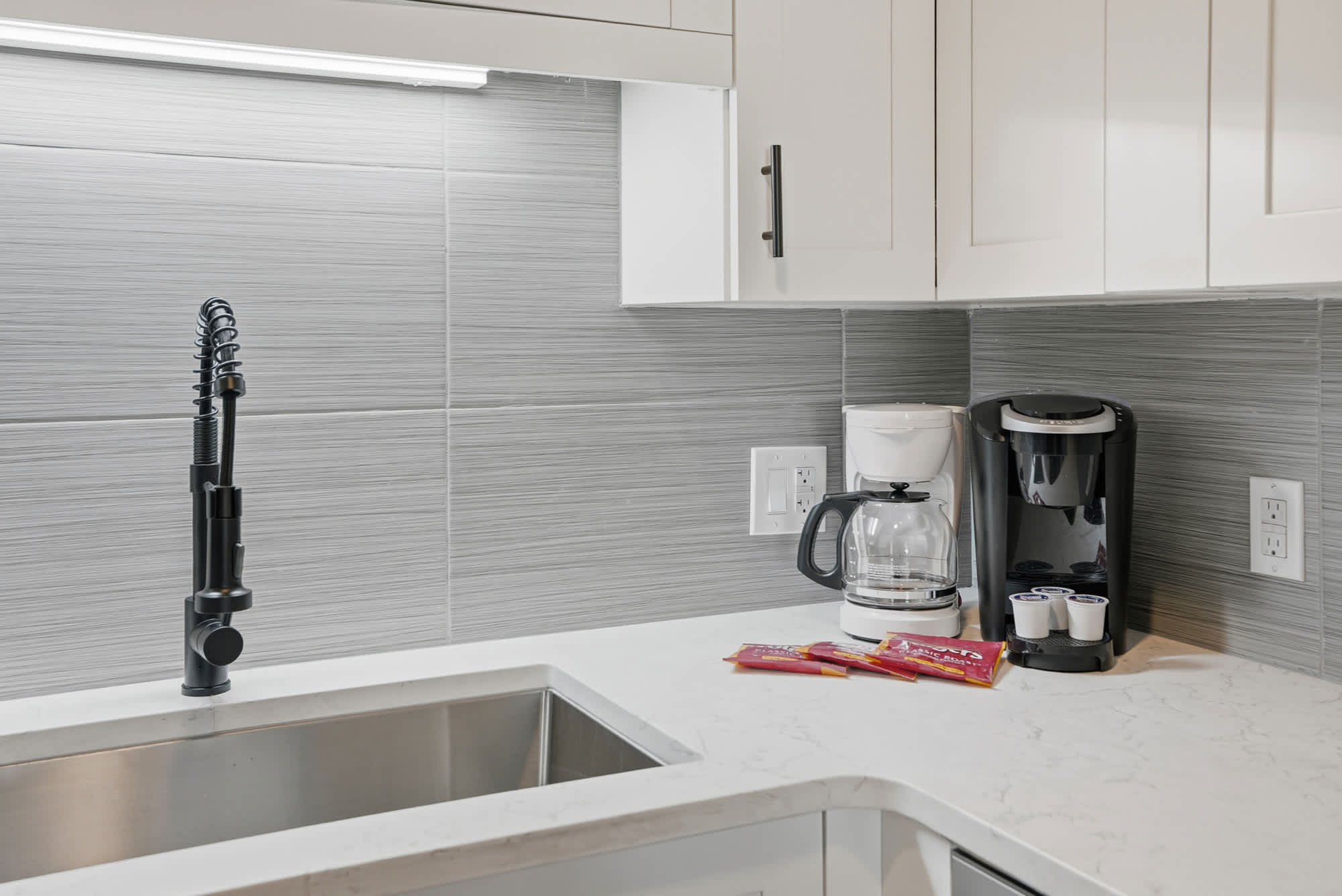 The Summit 1217 – Modern kitchen with stainless steel sink, black faucet, coffee maker, and snack packets on white countertop.
