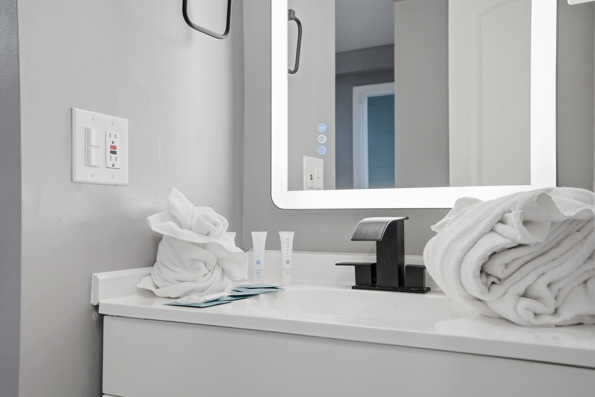 The Summit 1217 – Modern bathroom with a illuminated mirror, black faucet, towels, and complimentary toiletries on a white countertop.