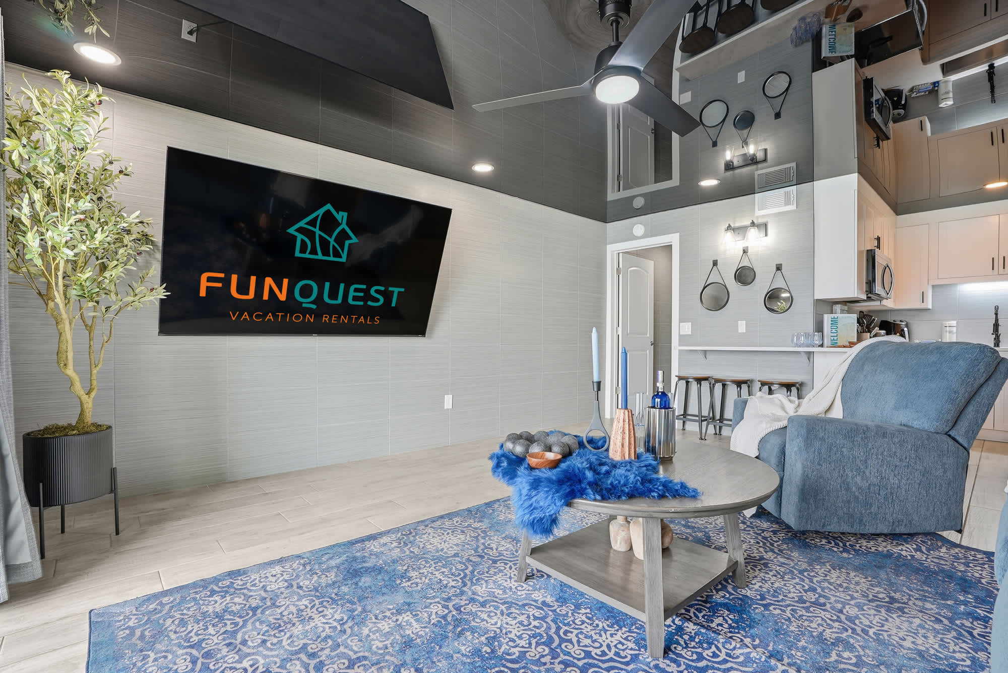 The Summit 1217 – Living room with TV, seating area, decorative plants, and kitchenette in background.