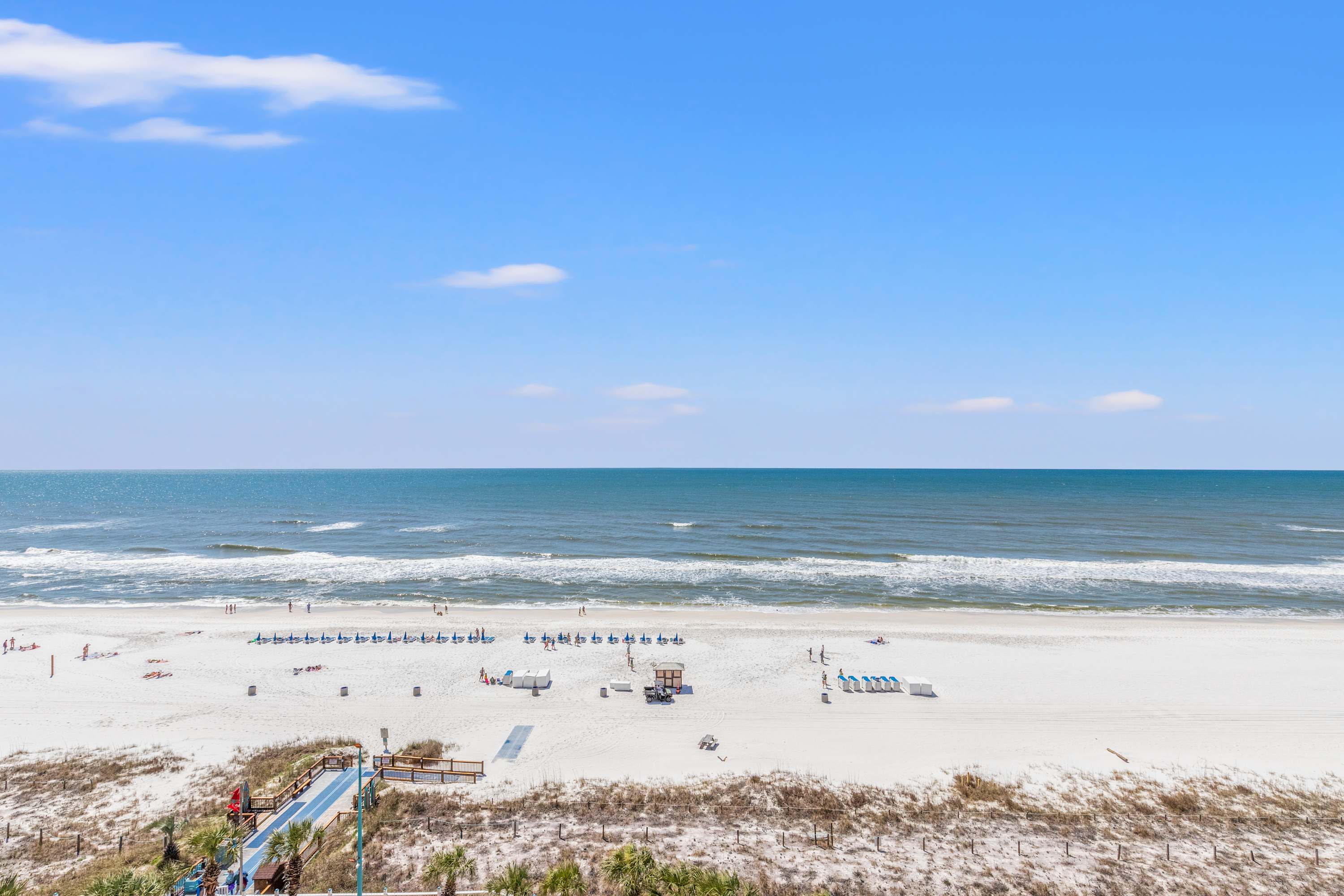 The Summit 1217 – View of a sandy beach with ocean waves and lounge chairs under a clear blue sky.