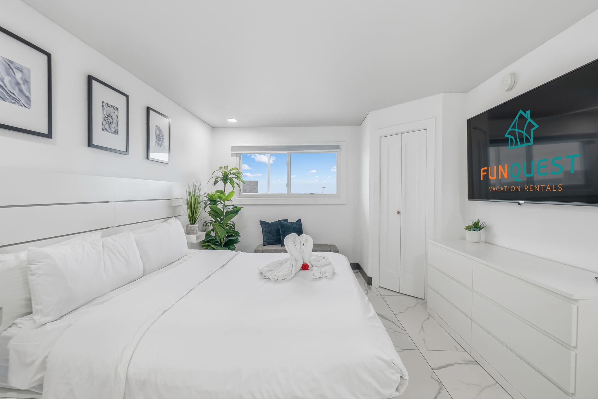 The Summit 1208 – Bedroom with a large bed, TV, sofa, plants, and a window showing a blue sky.