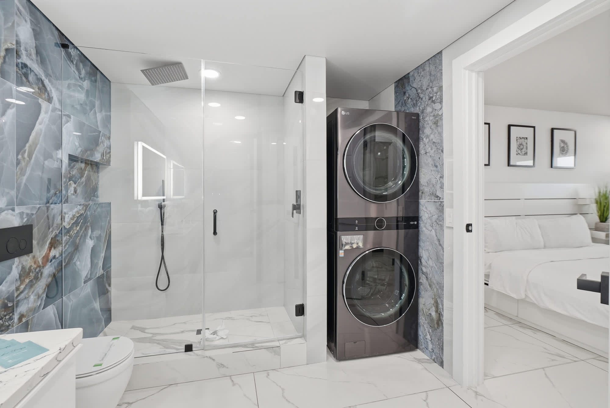 The Summit 1208 – Modern bathroom with glass shower, stacked washer and dryer, and a glimpse of a bedroom with white bedding.