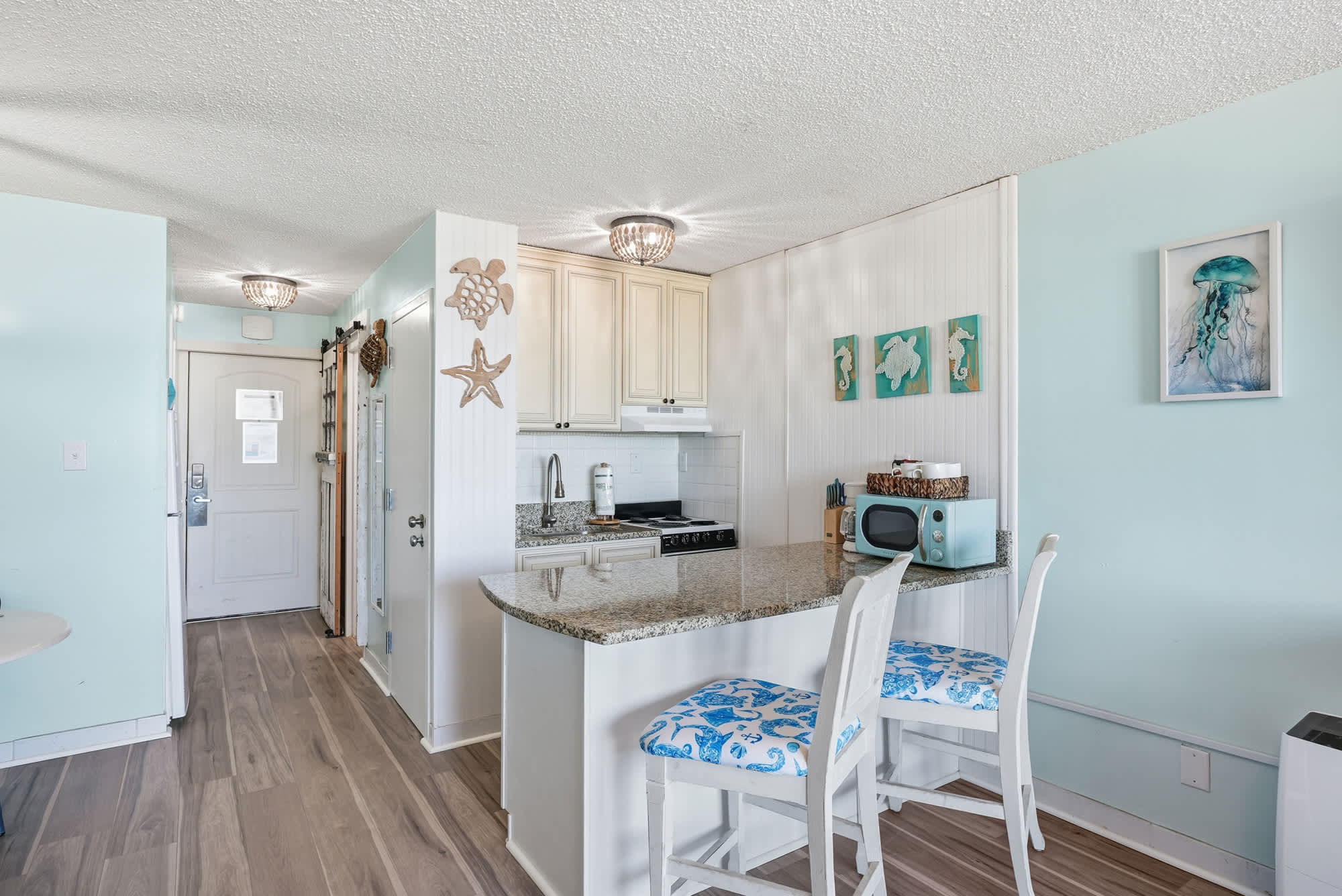 Top Of The Gulf  602 – Modern kitchen area with granite countertops, two bar stools, and light blue walls.