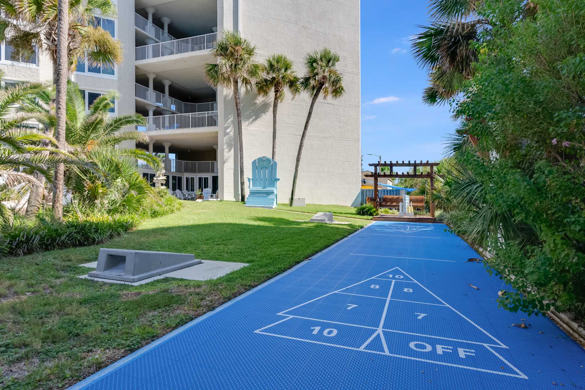 Top Of The Gulf 613 – Outdoor area with shuffleboard court, palm trees, and seating near a multi-story rental building.