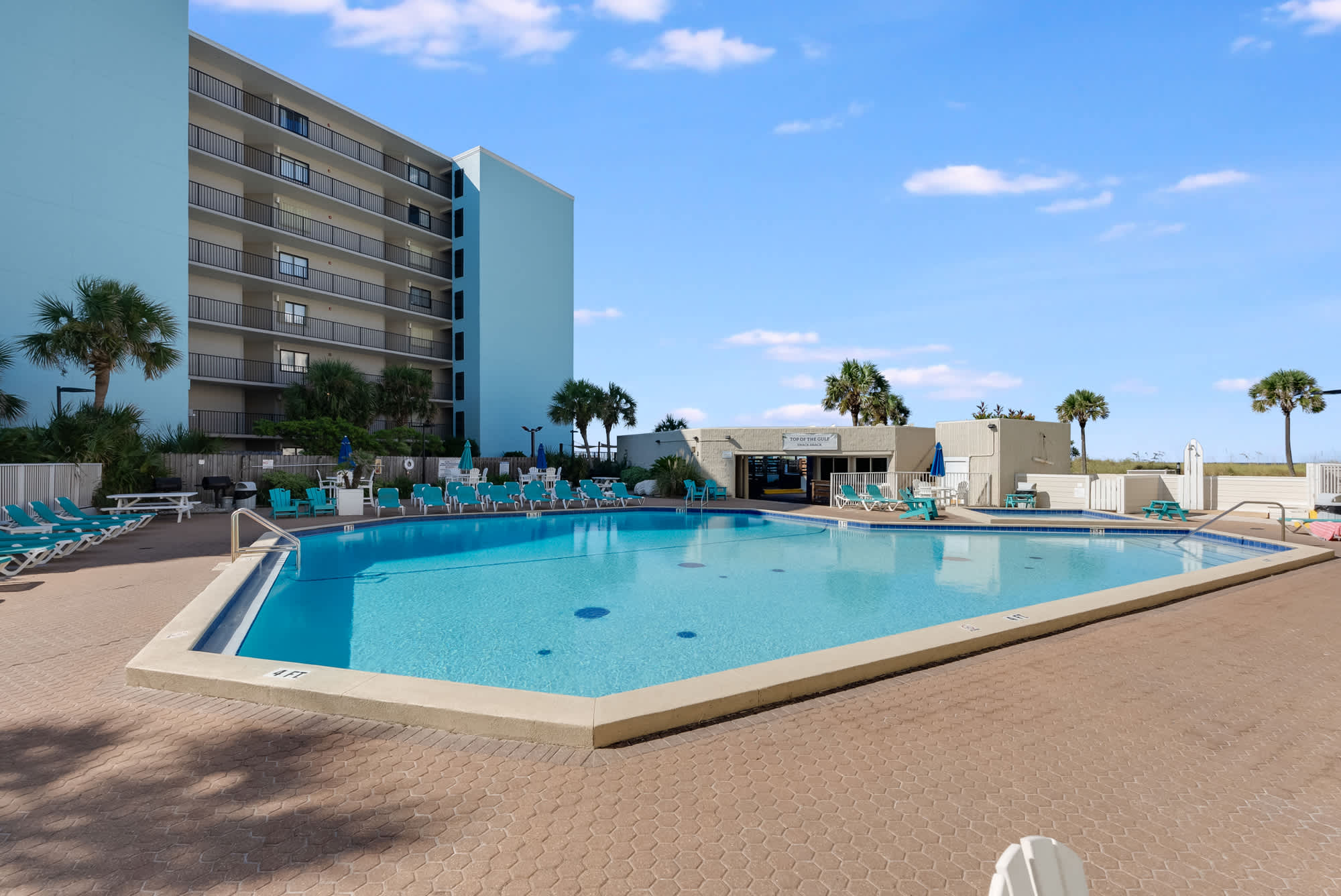 Top Of The Gulf 613 – Swimming pool area with lounge chairs and a multi-story blue building in the background.