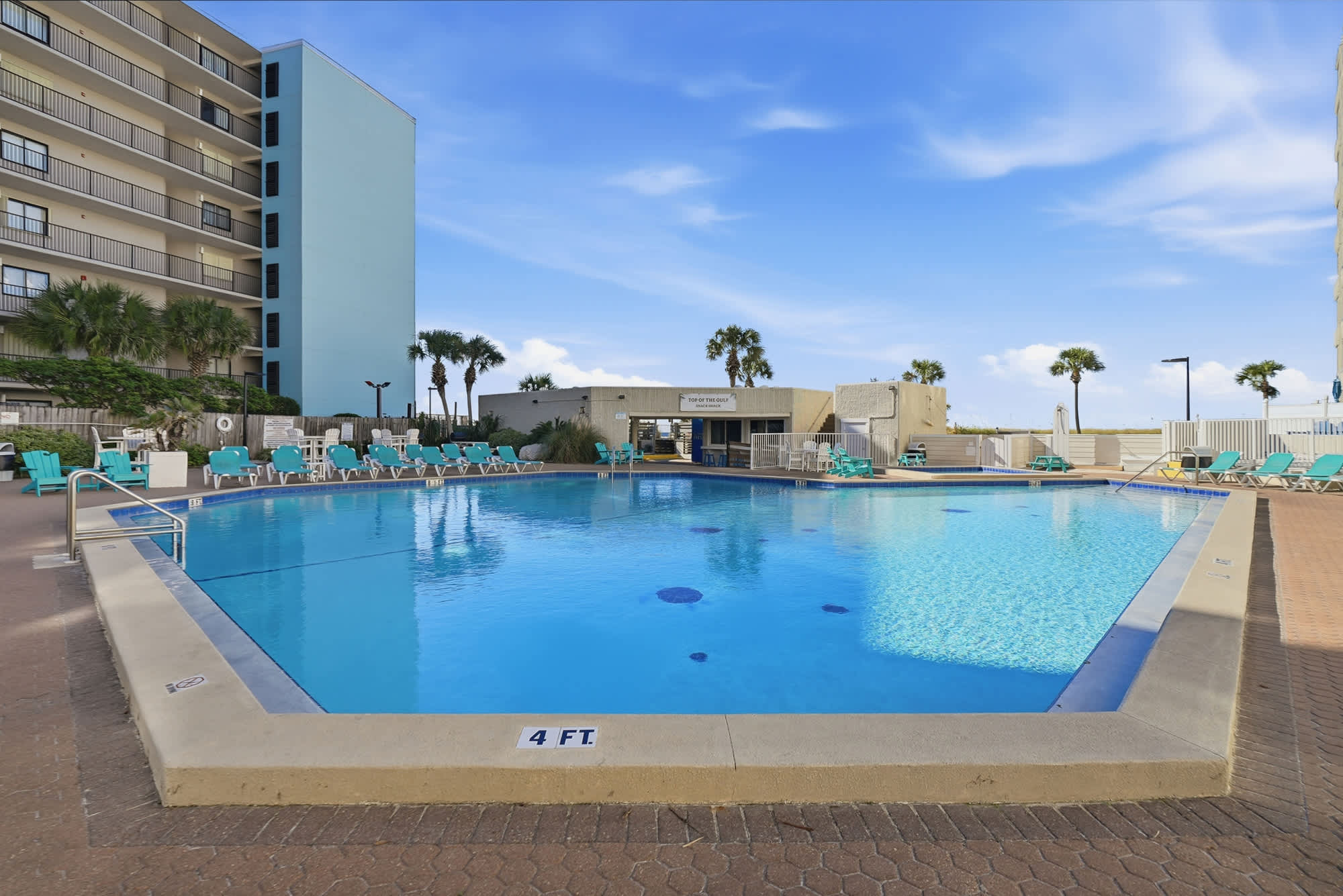 Top Of The Gulf 704 – Swimming pool area with lounge chairs and a building in the background, under a clear blue sky.