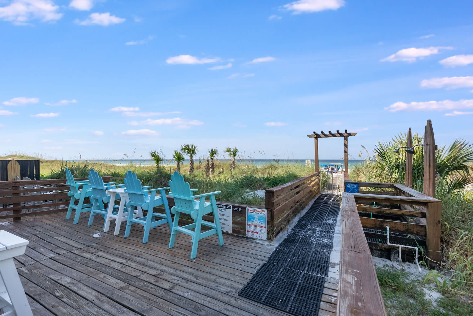 Top Of The Gulf 704 – Wooden deck with blue chairs, leading to beach access through dunes and palm trees.