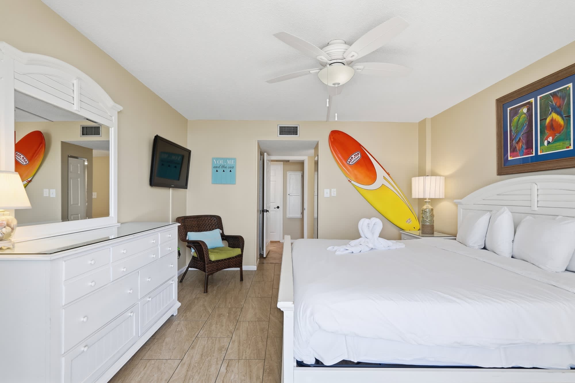 Dunes C104 – Cozy bedroom with white furniture, surfboard decor, flat-screen TV, and artwork on the walls.