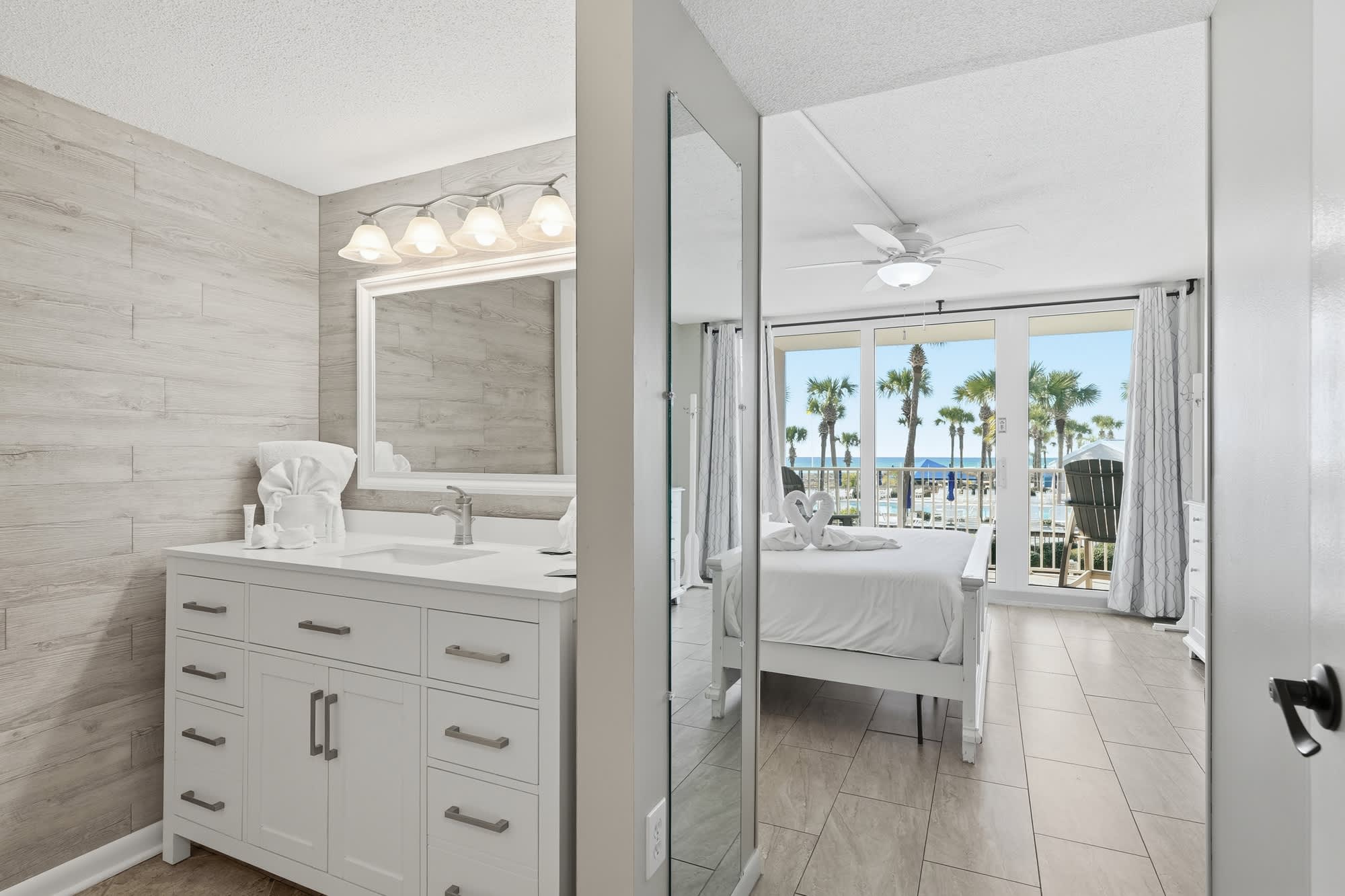 Dunes C104 – Bright bedroom with queen bed, adjacent bathroom, and large windows showing palm trees and ocean view.