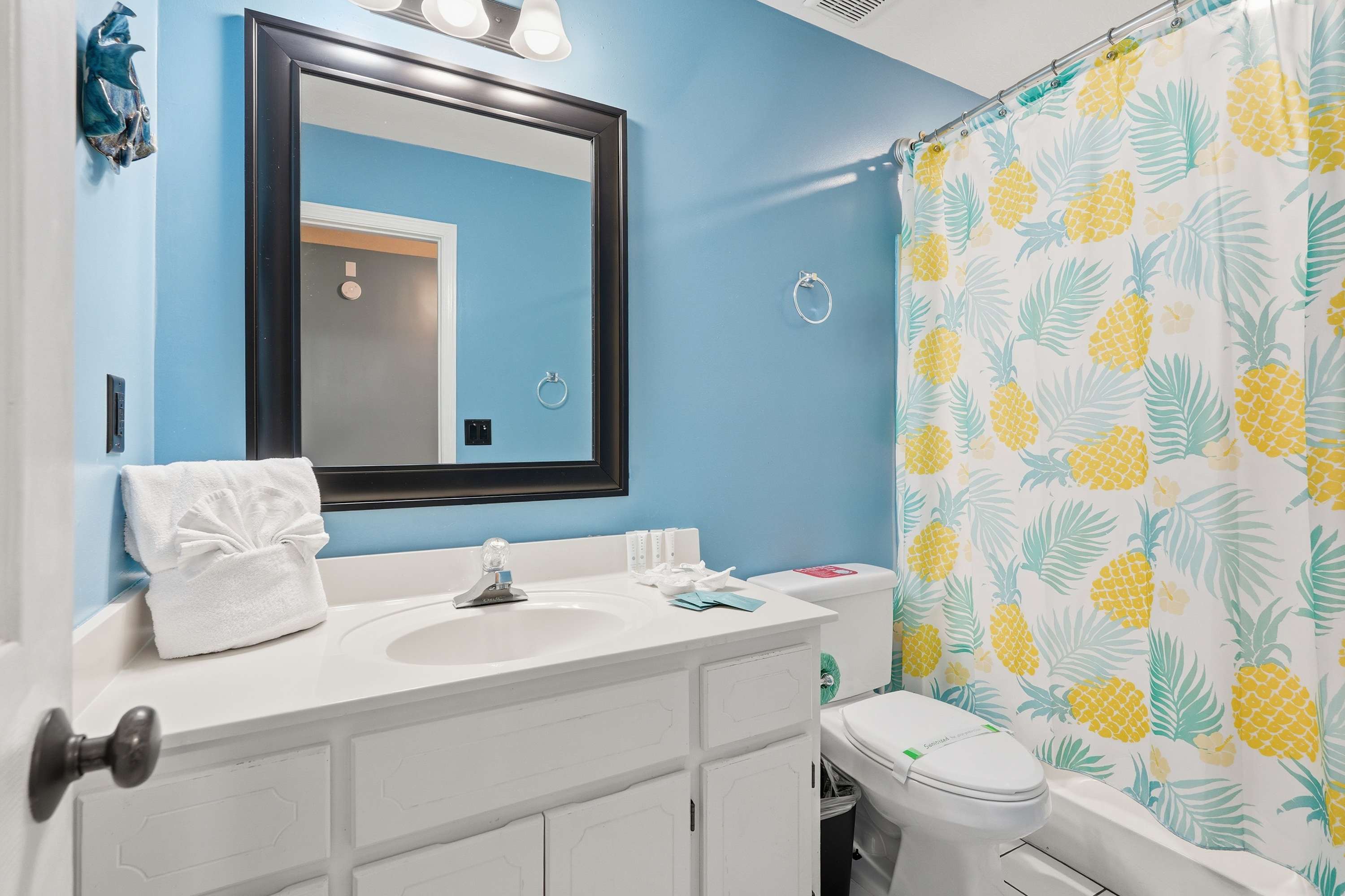 135 Grande Island Blvd – Bright bathroom with blue walls, white vanity, large mirror, and pineapple-patterned shower curtain.