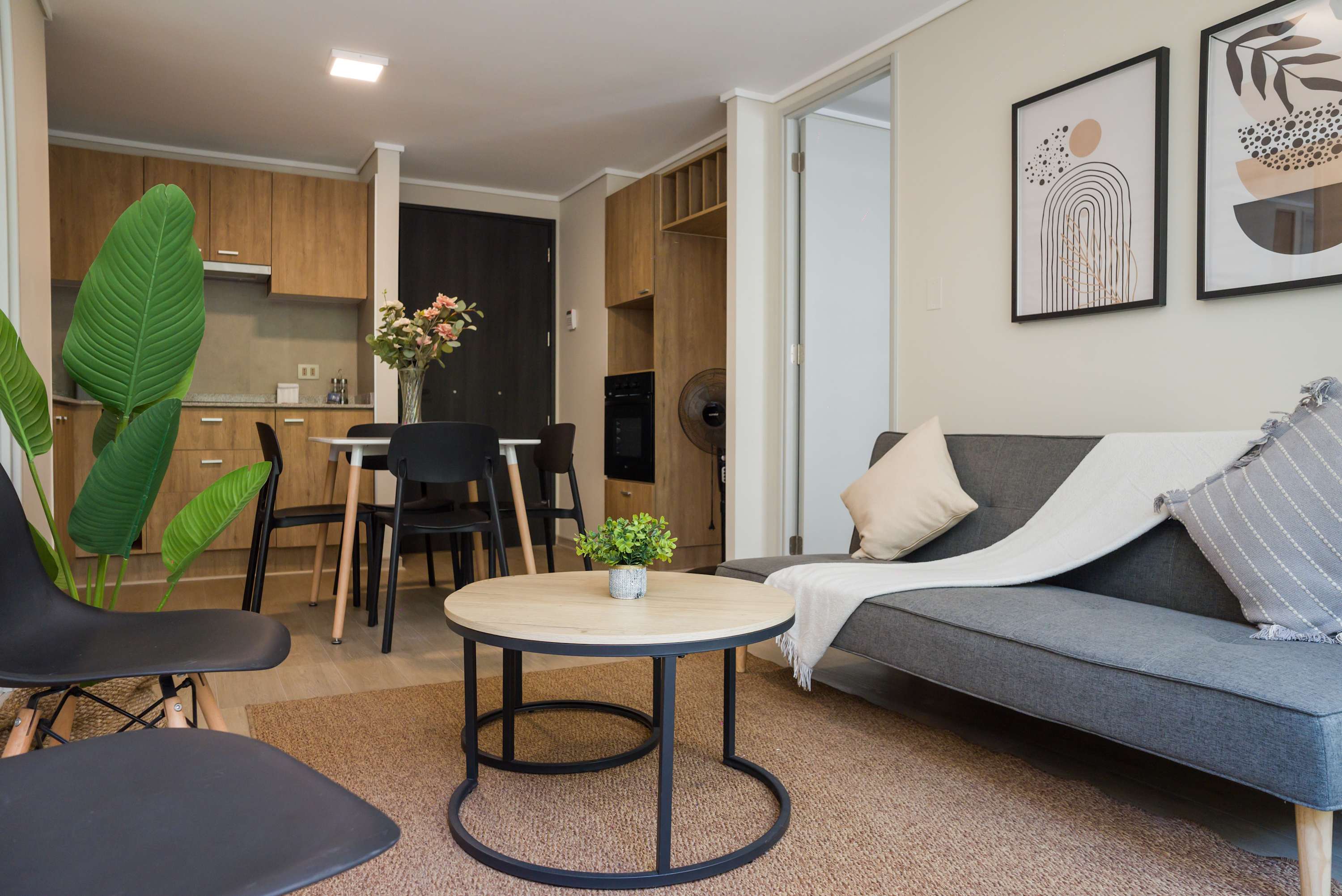 Open-plan living room with a gray sectional, textured throw, and round coffee tables beside a wood dining and kitchen area.
