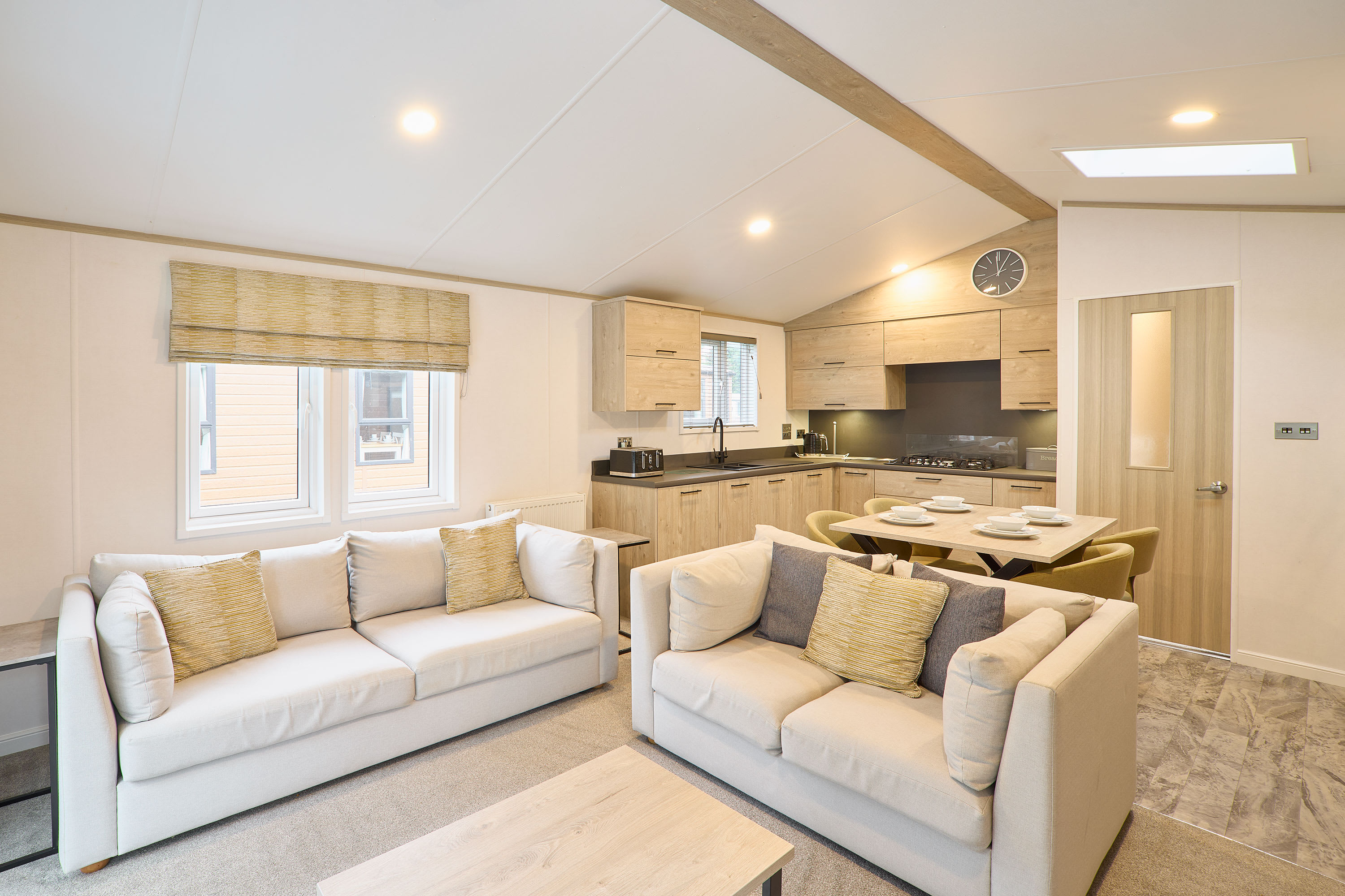 Open-plan living space with two light-colored sofas, a wooden coffee table, and a view into the modern kitchen and dining area with light wood cabinetry and a skylight.