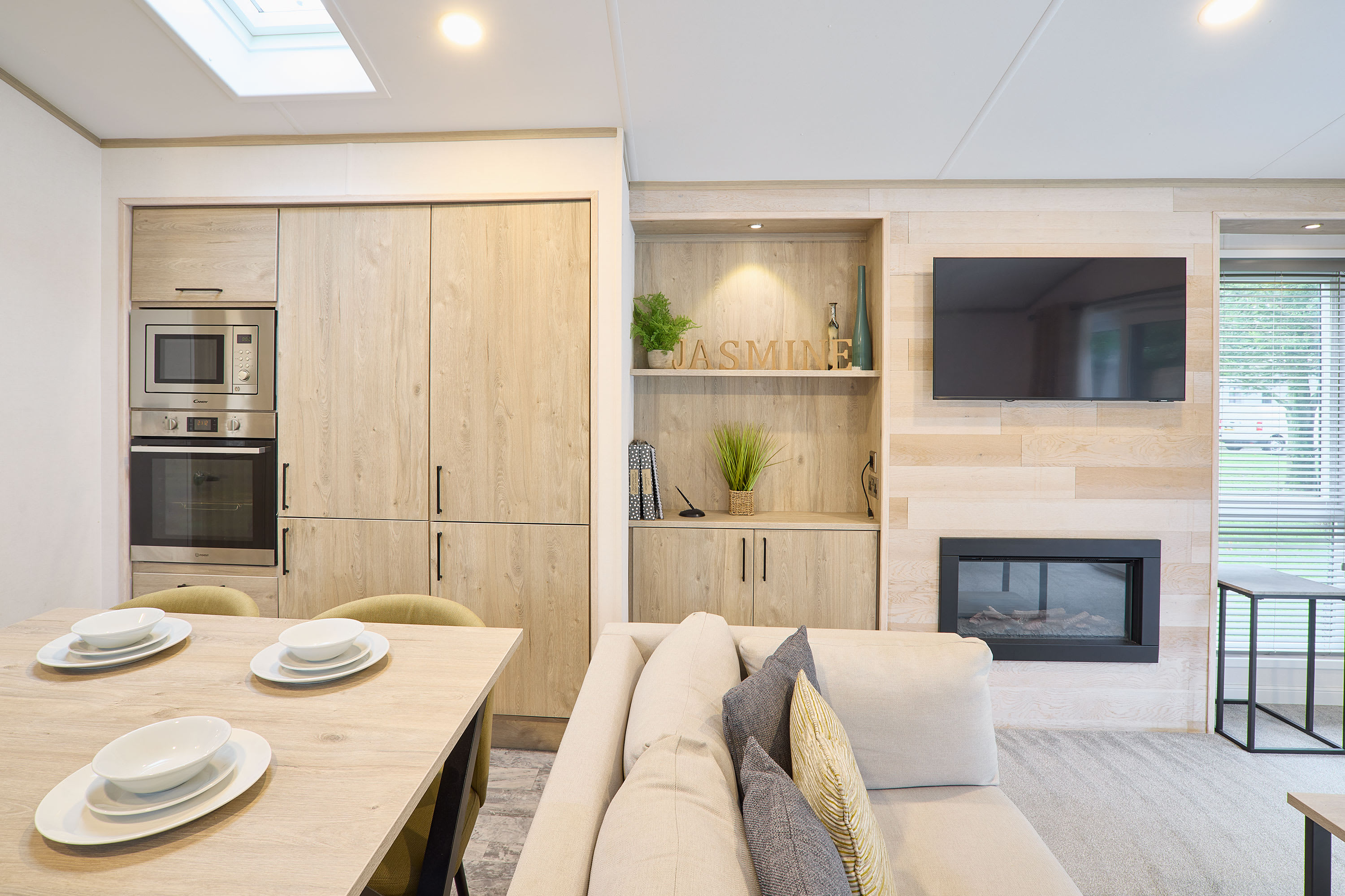 Expansive open-plan living area featuring a light wood kitchen with integrated appliances, a dining table with four chairs, and a comfortable sofa. A wall-mounted TV and fireplace are focal points in the living space.