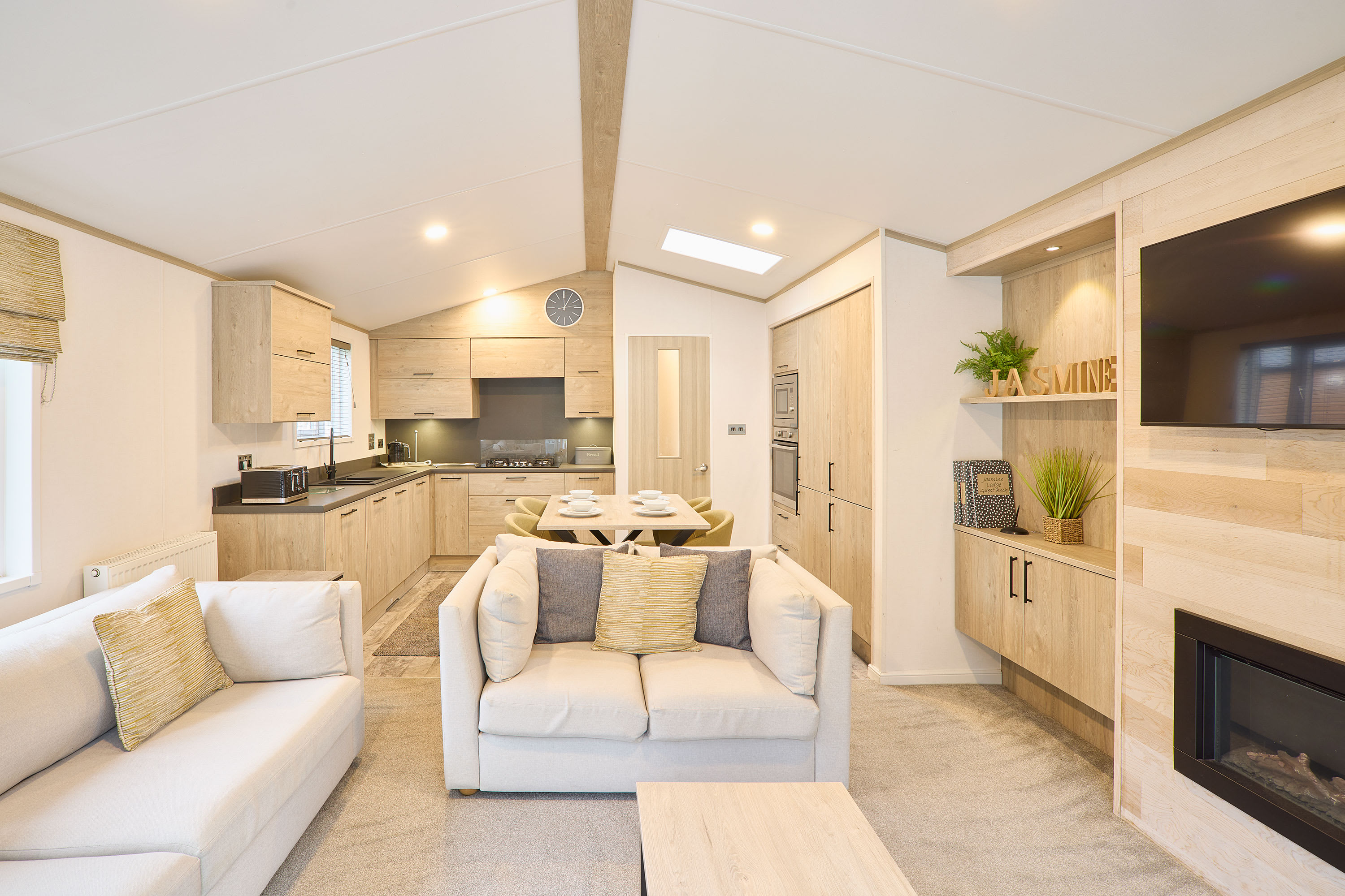 Bright open-plan living area featuring two light sofas, a coffee table, a wall-mounted TV above a fireplace, and an integrated kitchen and dining space with light wood finishes.