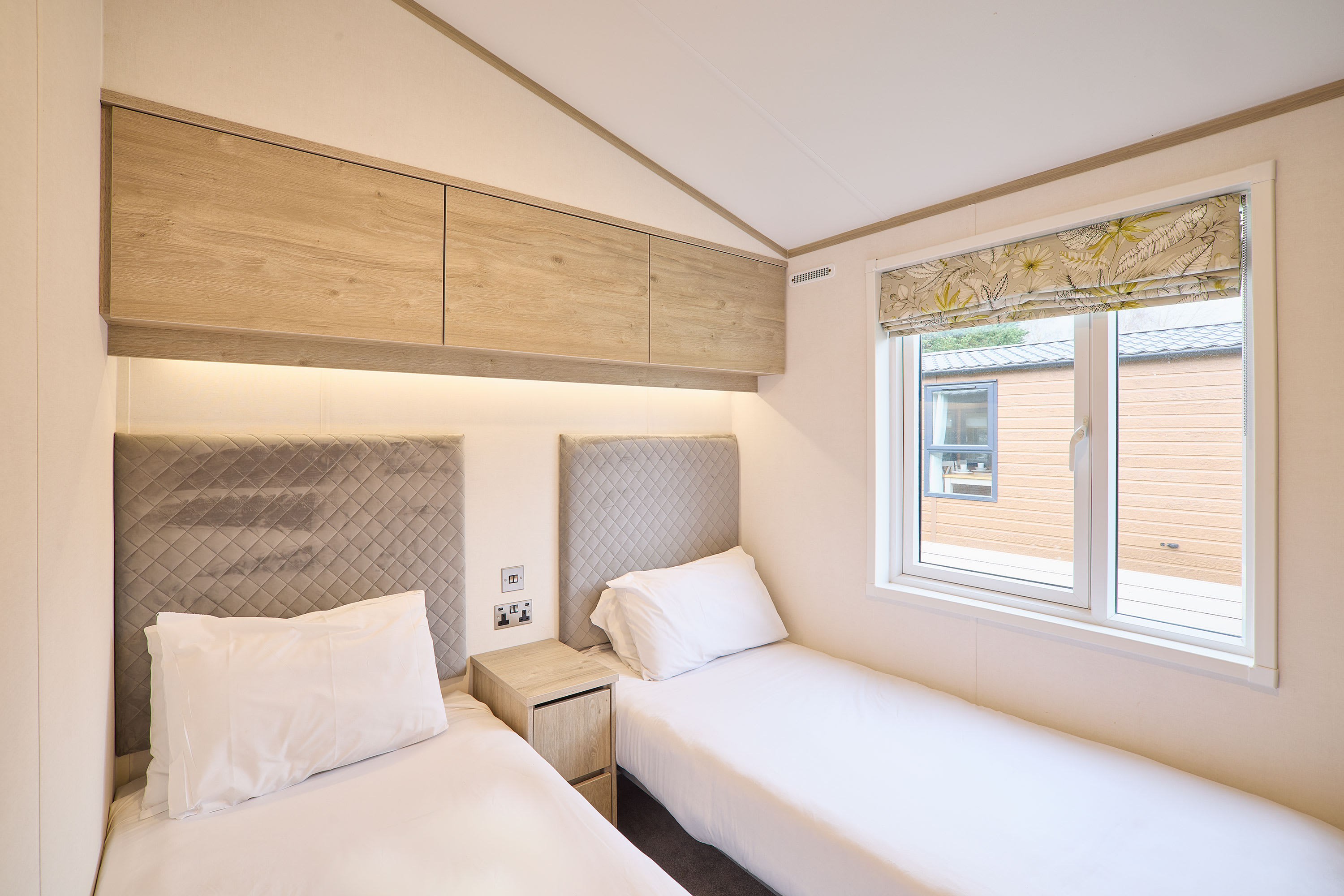 Another view of the twin bedroom, showing two single beds with upholstered headboards, a central nightstand, and light wood overhead cabinets, illuminated by natural light from a large window.