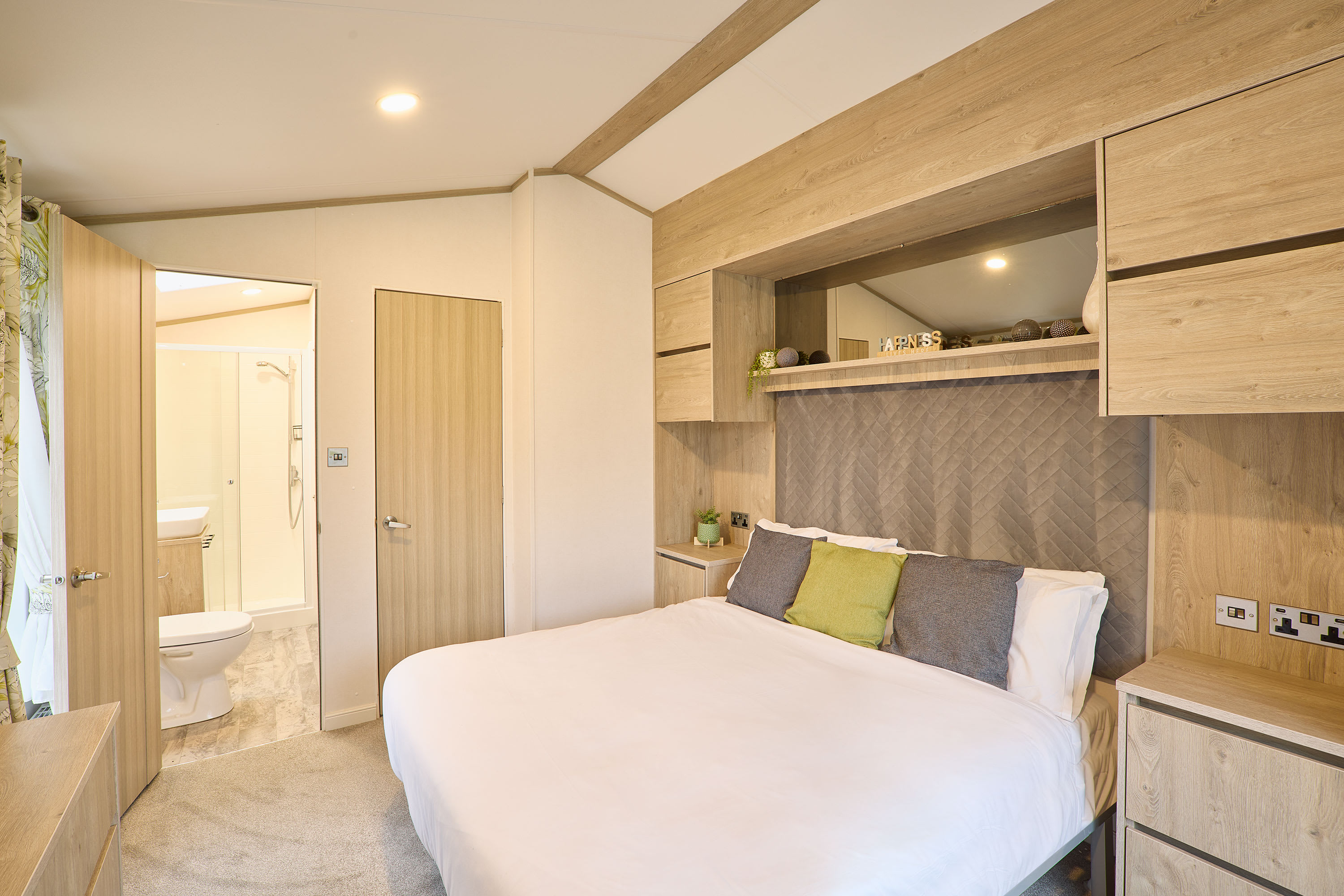 Master bedroom with a double bed, upholstered headboard, built-in light wood storage above and beside the bed, and an en-suite bathroom visible through an open door.