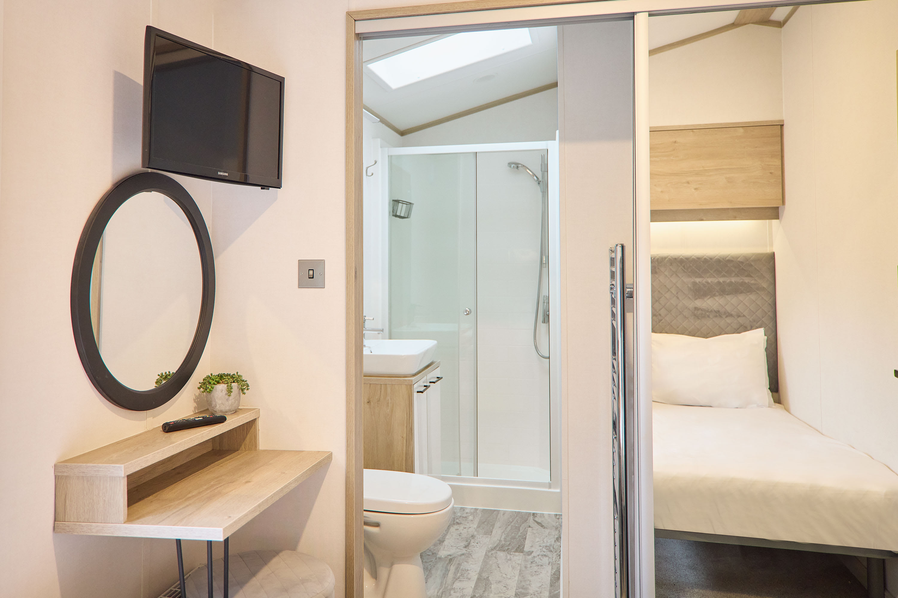 Compact sleeping area with a single bed, a vanity desk with a round mirror, and a wall-mounted TV, with a modern bathroom featuring a shower and toilet visible through a sliding glass door.