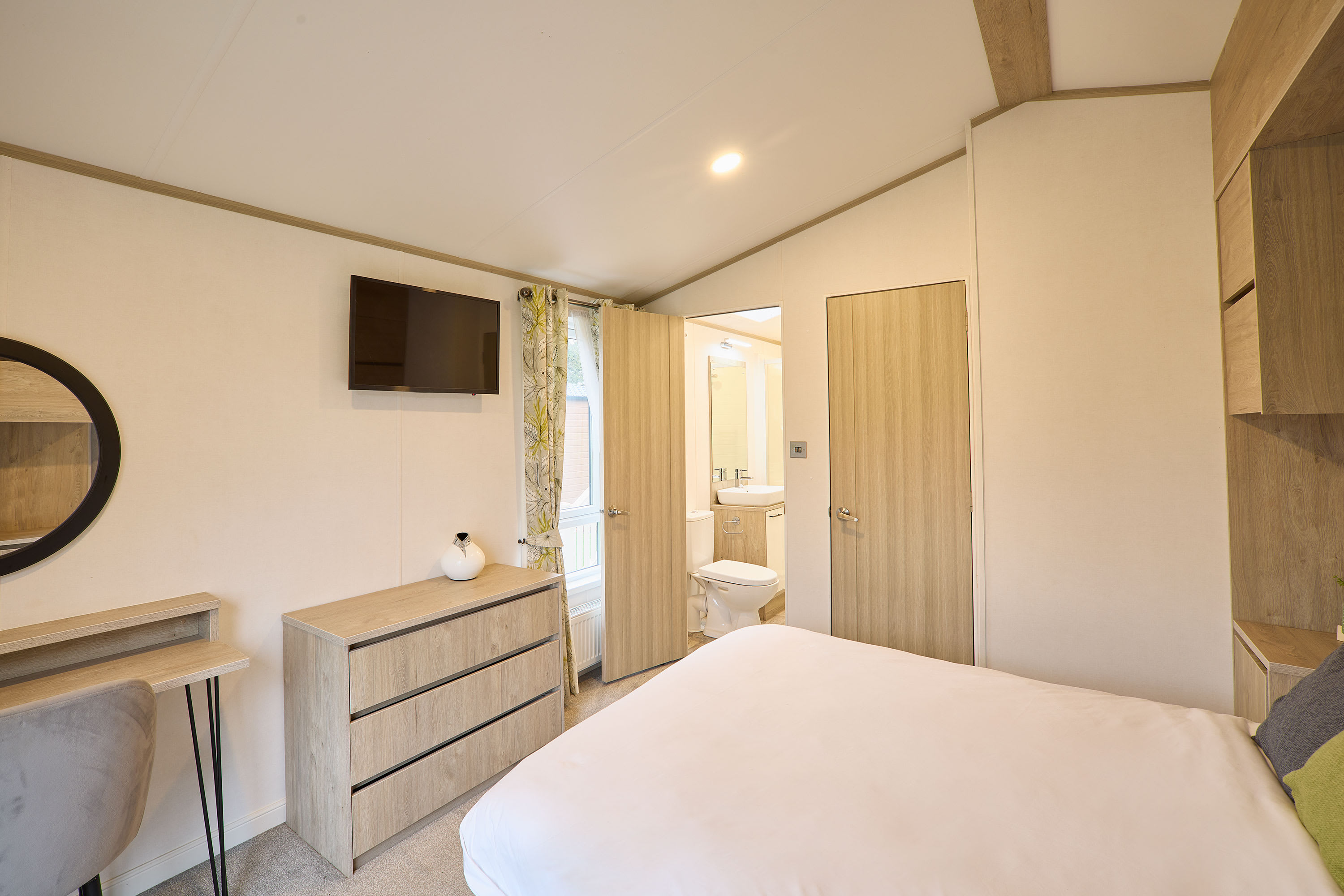 Cozy bedroom with a double bed, light wood dresser, vanity desk with a round mirror, wall-mounted TV, and an en-suite bathroom visible through an open door.