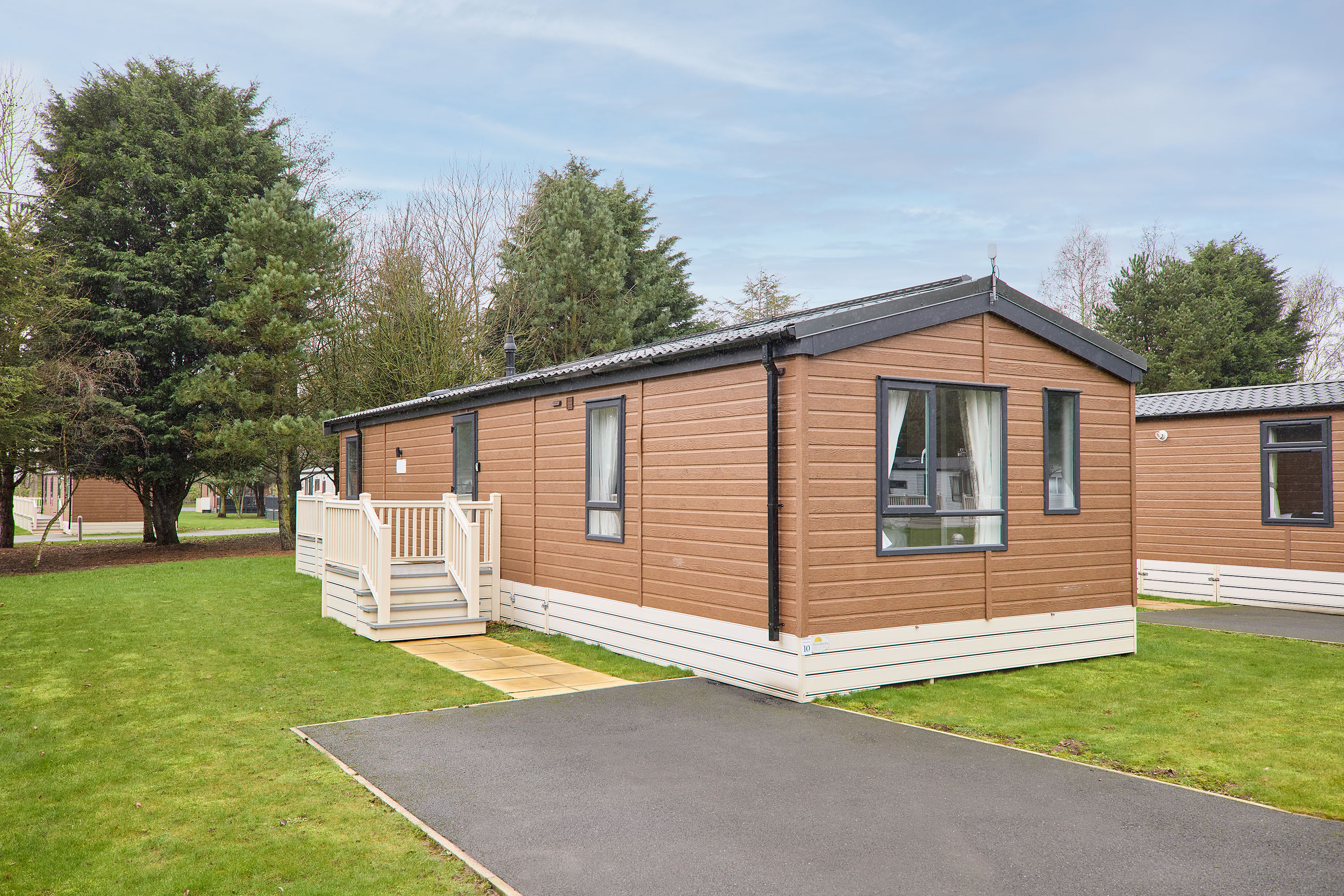 A modern, brown-sided chalet with black trim and white skirting, featuring multiple windows and a small white deck with steps leading to a paved driveway. It's set on a green lawn with evergreen trees in the background under a cloudy sky.